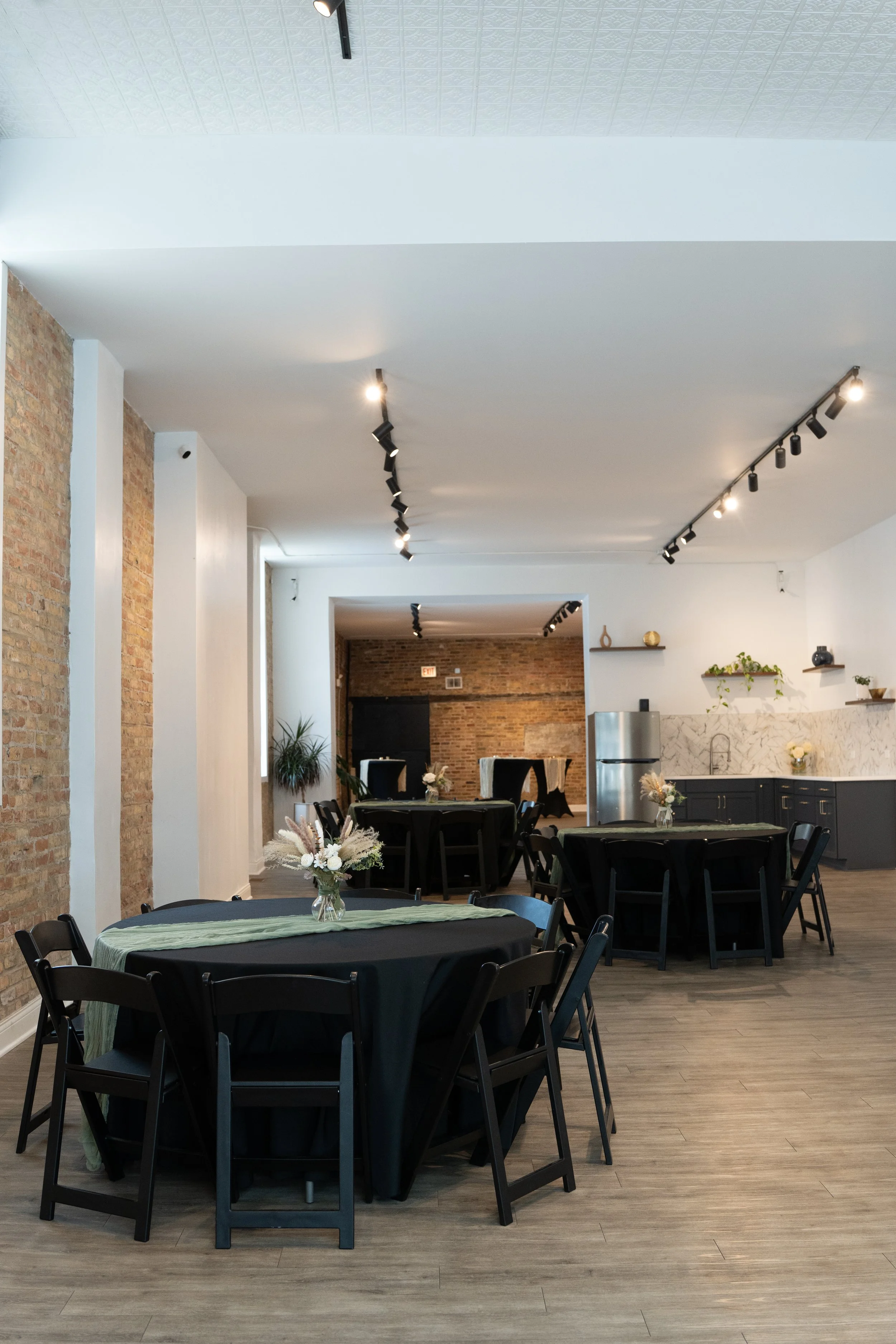 Event space with round tables covered with black tablecloths and light green table runners, decorated with floral centerpieces, in a room with exposed brick walls, white walls, and wooden flooring, with track lighting on the ceiling.