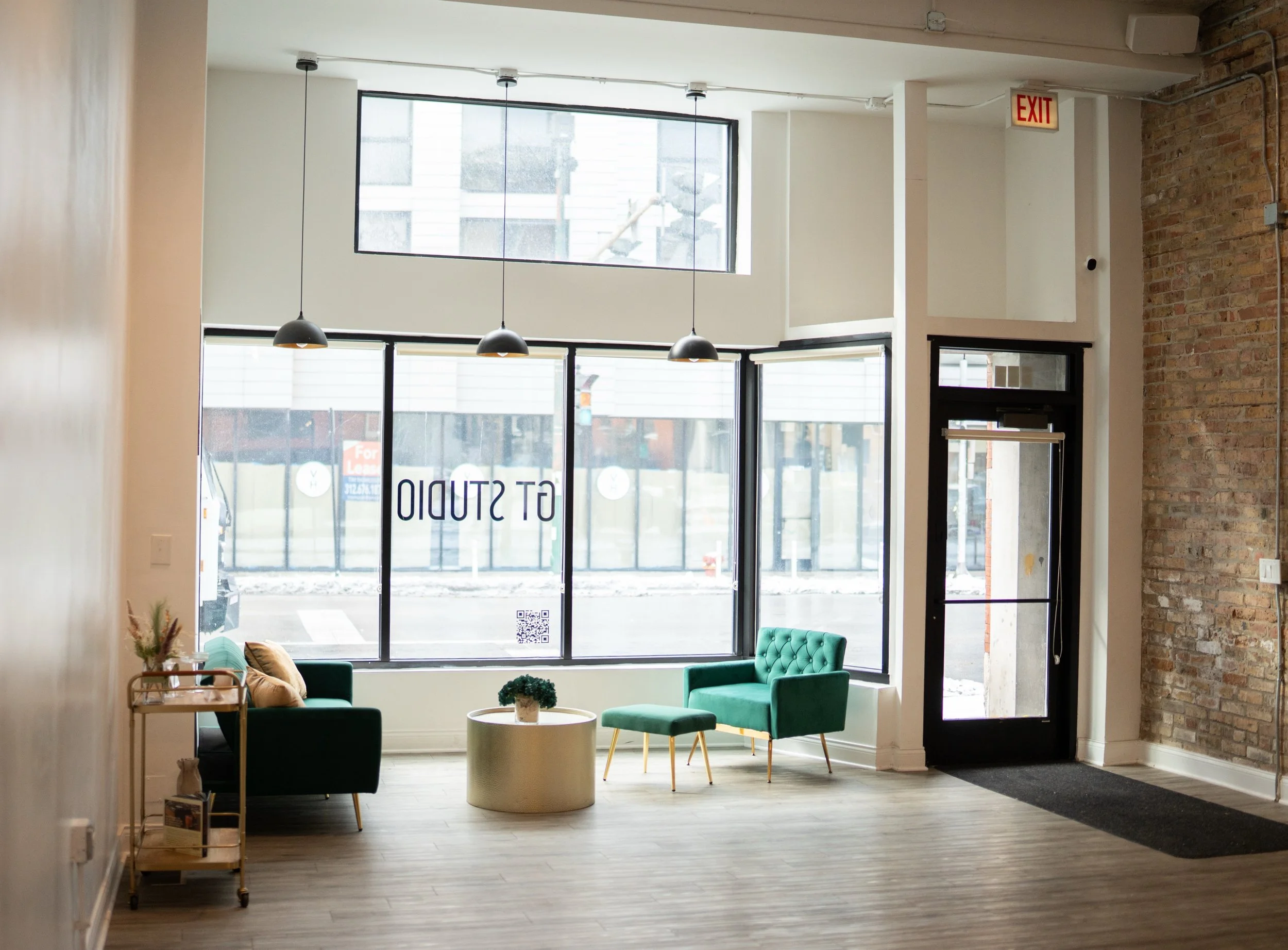 Interior of a modern building with large windows, hanging pendant lights, green sofas, a round coffee table with a plant, and brick and white walls, indicating a cozy lounge or waiting area.