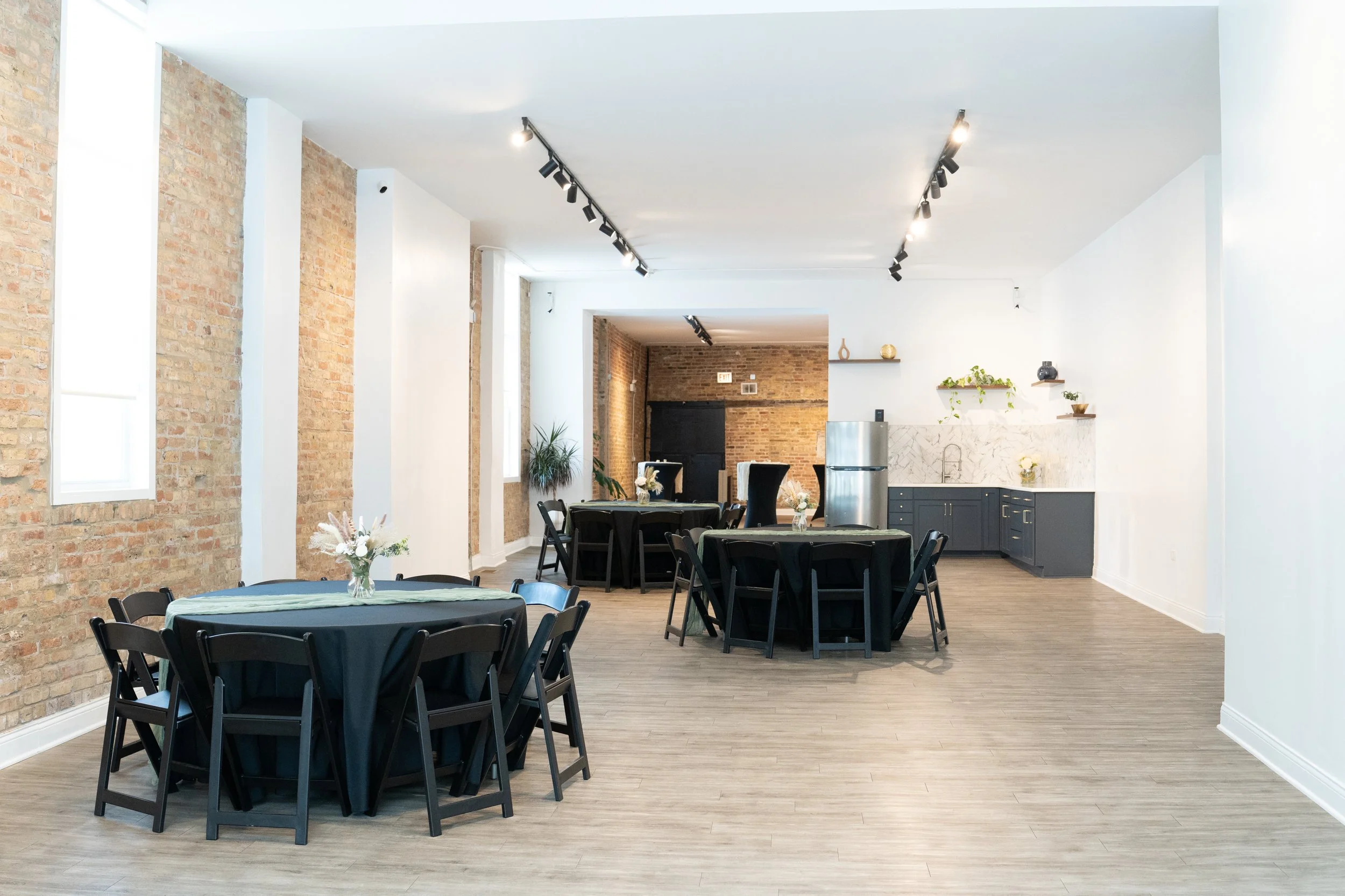 Interior of a modern event space with exposed brick walls, white walls, track lighting, and round tables with black chairs and floral centerpieces.