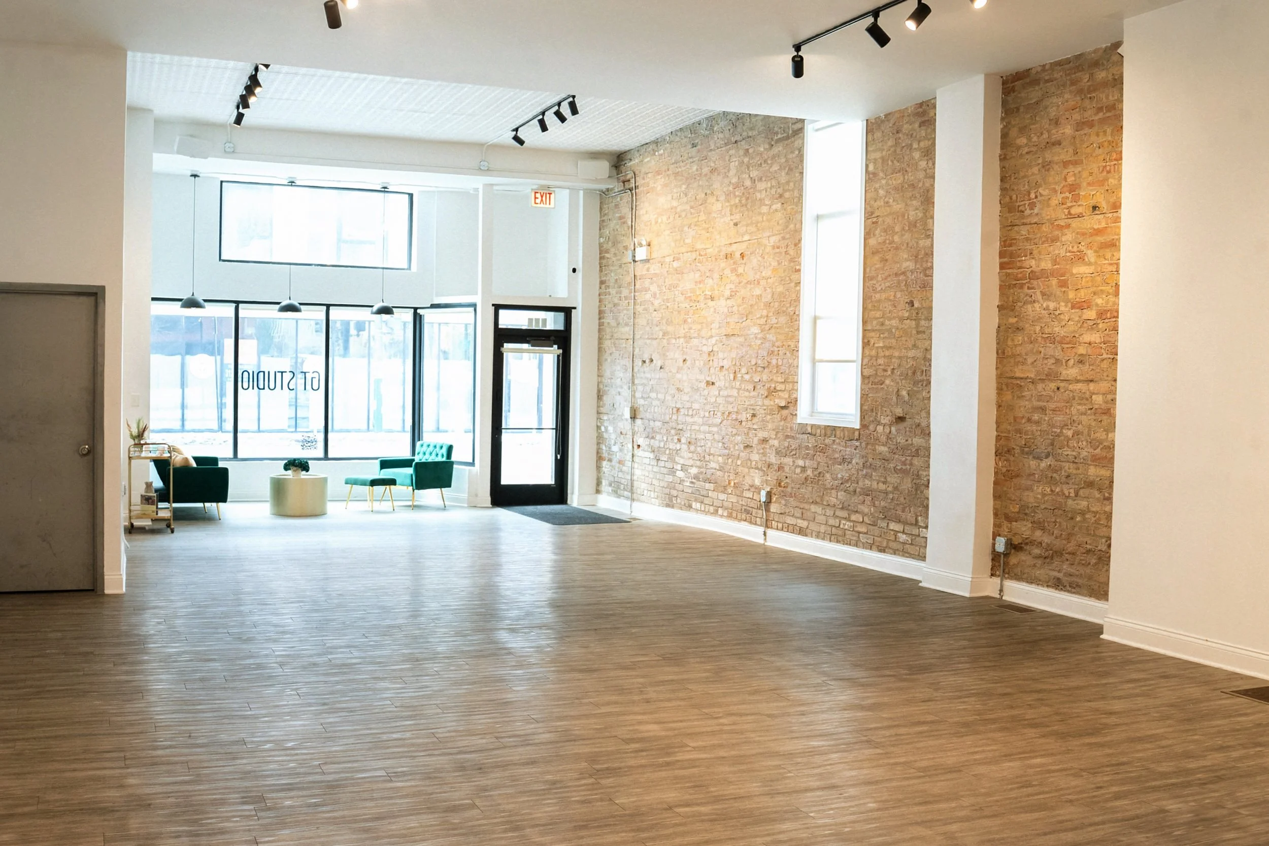 Interior of a modern space with a brick wall, large windows, and bamboo flooring, featuring green chairs and a small table near the window.