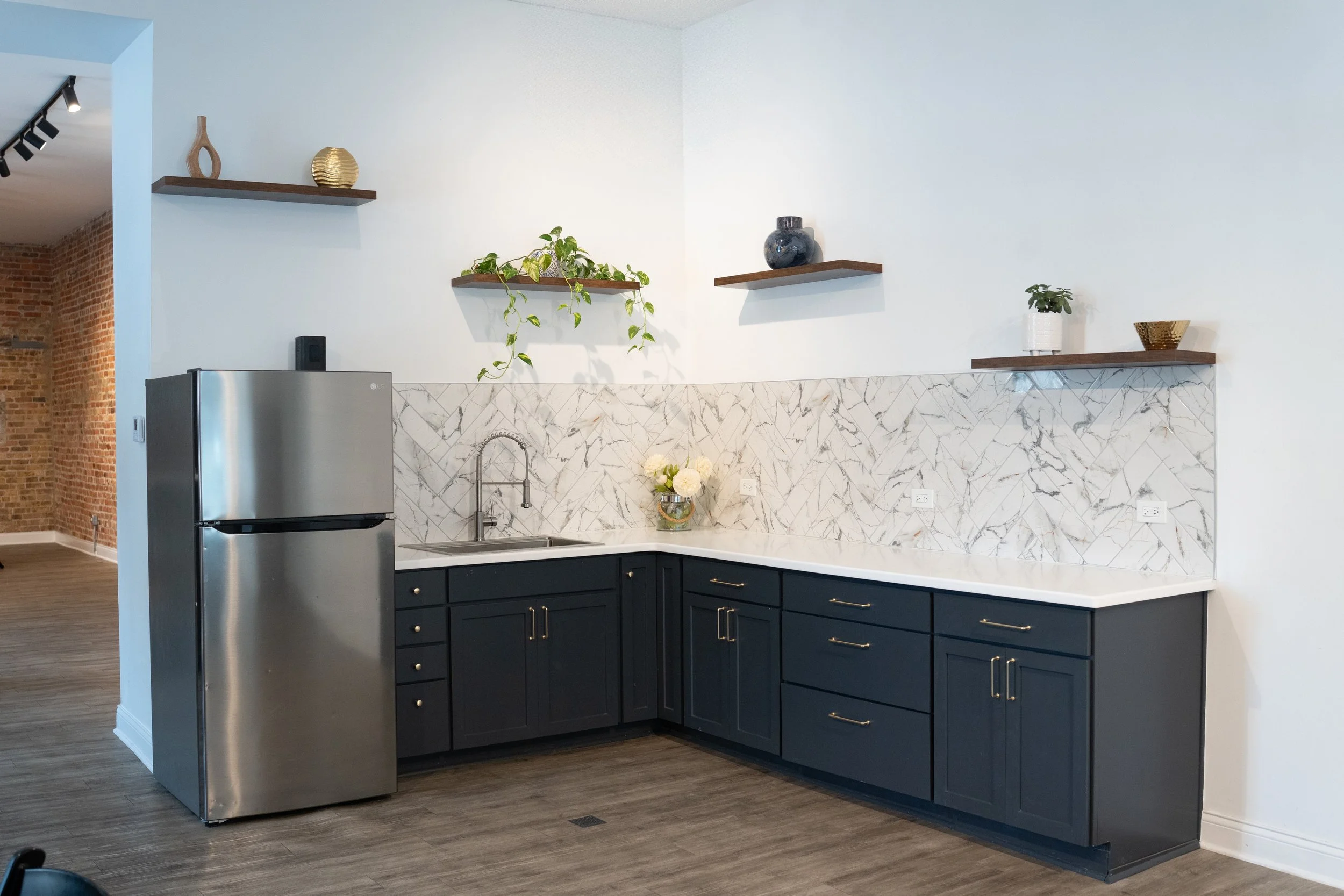 Modern kitchen with black cabinets, marble backsplash, stainless steel refrigerator, decorative shelves with vases and plants, and wooden flooring.