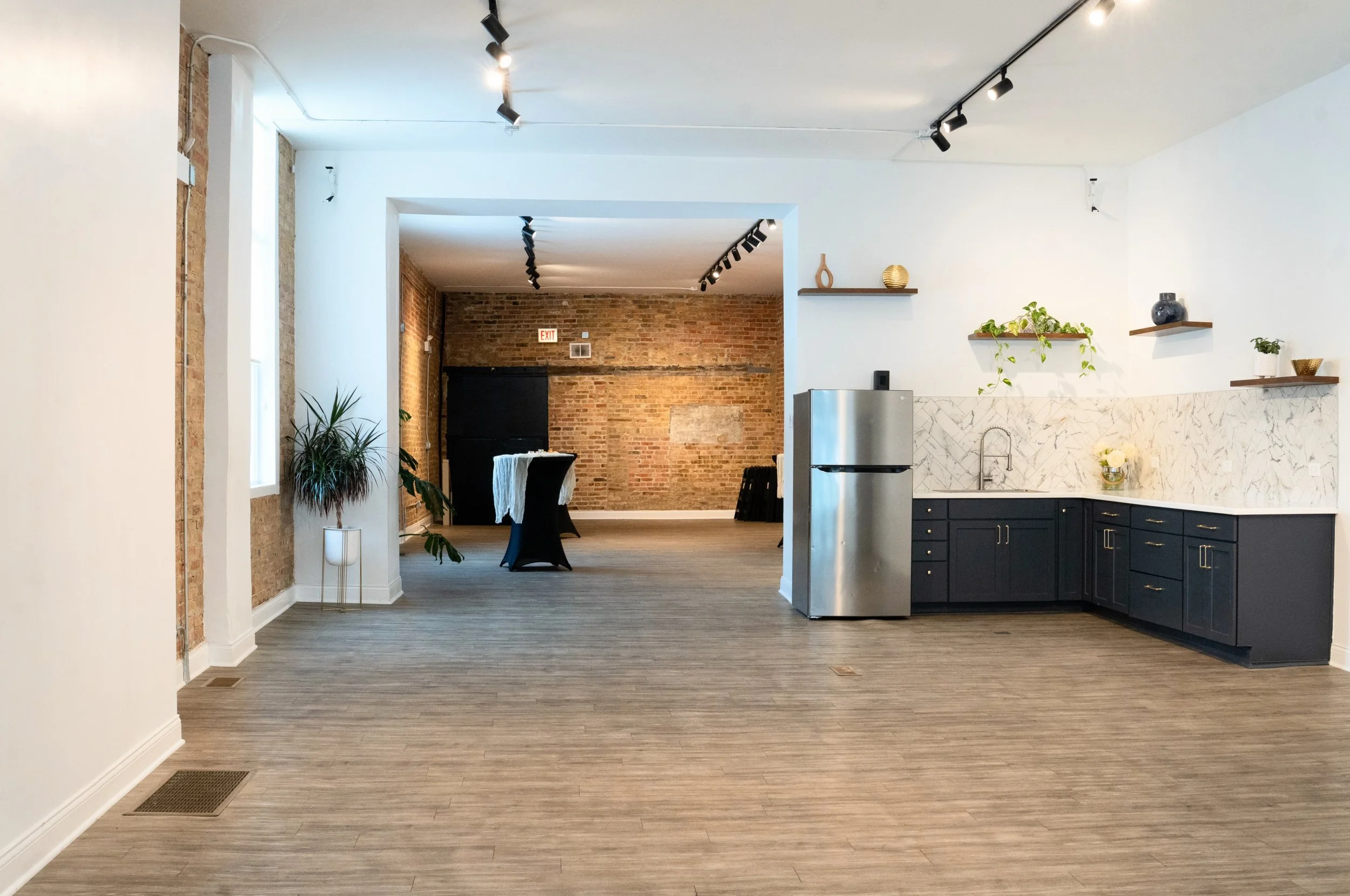 An open-concept kitchen and dining area with exposed brick walls, white and navy cabinetry, and wooden shelves decorated with vases and plants. A small refrigerator is visible near the kitchen counter. The space has track lighting and wooden flooring