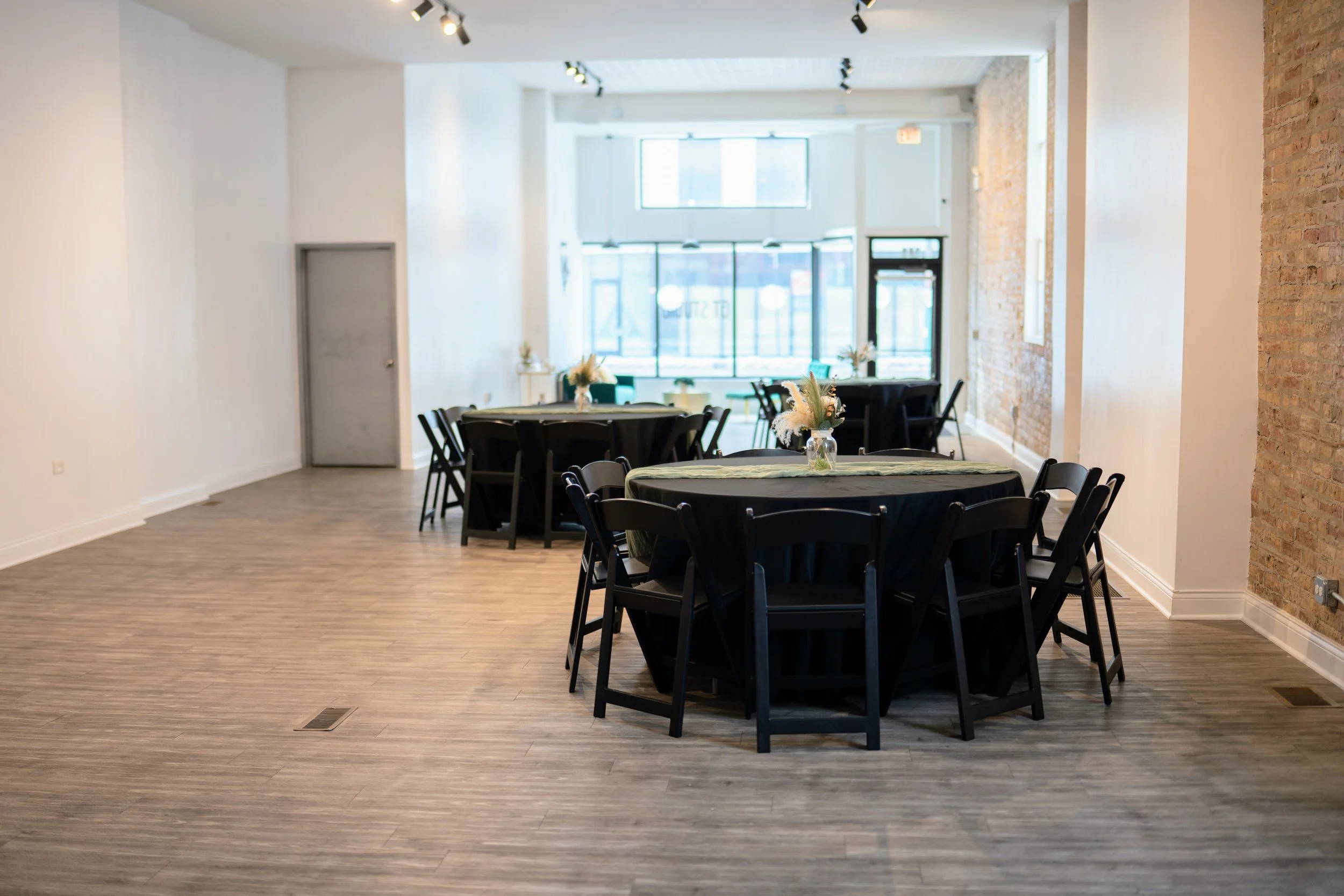 Interior of a bright, modern event space with round tables covered in dark tablecloths and floral centerpieces, large windows, exposed brick wall, and wooden floors.