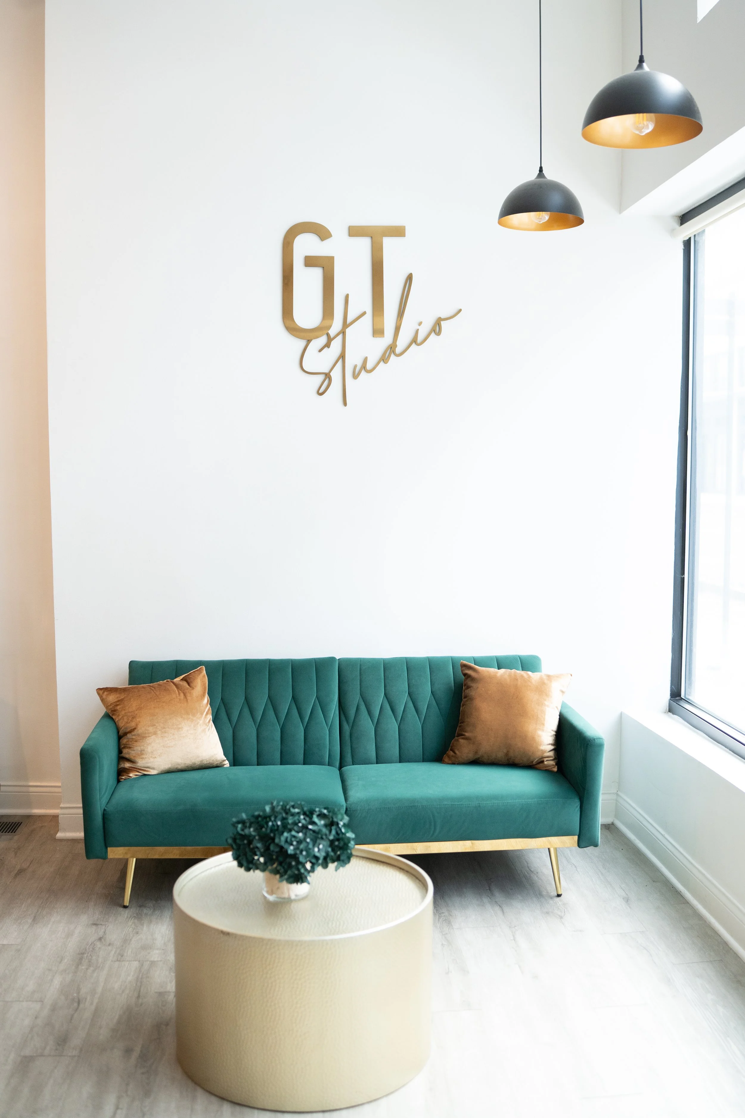 Modern coworking space with a teal velvet sofa, beige pillows, a white round table with a plant, a white wall with a gold sign that says 'GT Studio', black pendant lights, and a large window with natural light.