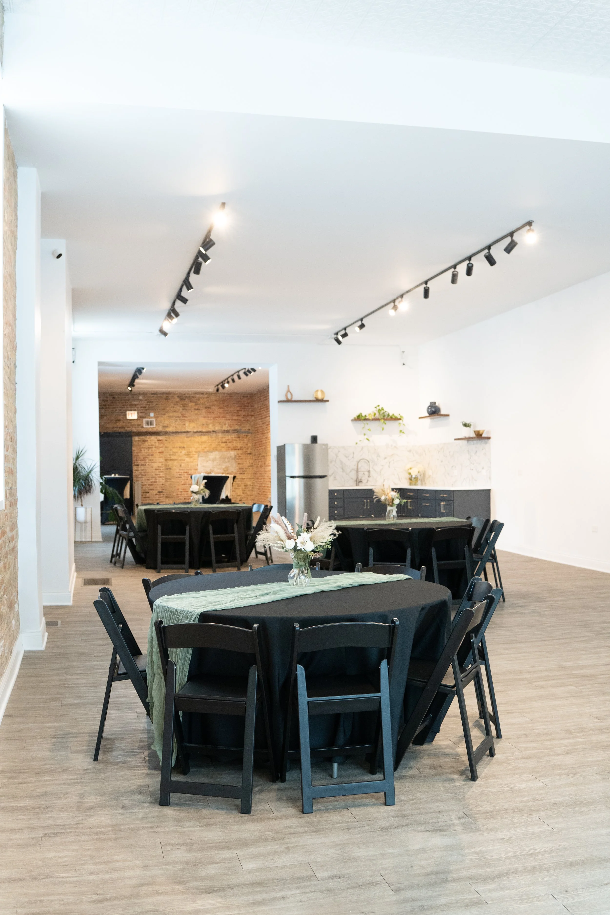 Elegant dining area with black tables covered with black tablecloths and decorated with small floral centerpieces, surrounded by black chairs, in a modern space with white walls, wooden flooring, track lighting on the ceiling, and a bar area with she