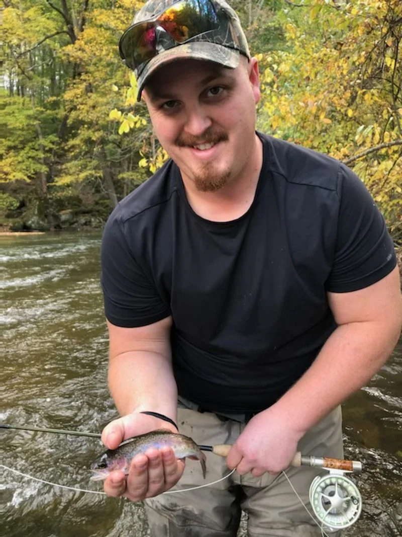 A young man wearing a camouflage cap, sunglasses, and a black t-shirt, smiling while holding a small fish he caught in a river surrounded by trees with autumn foliage.
