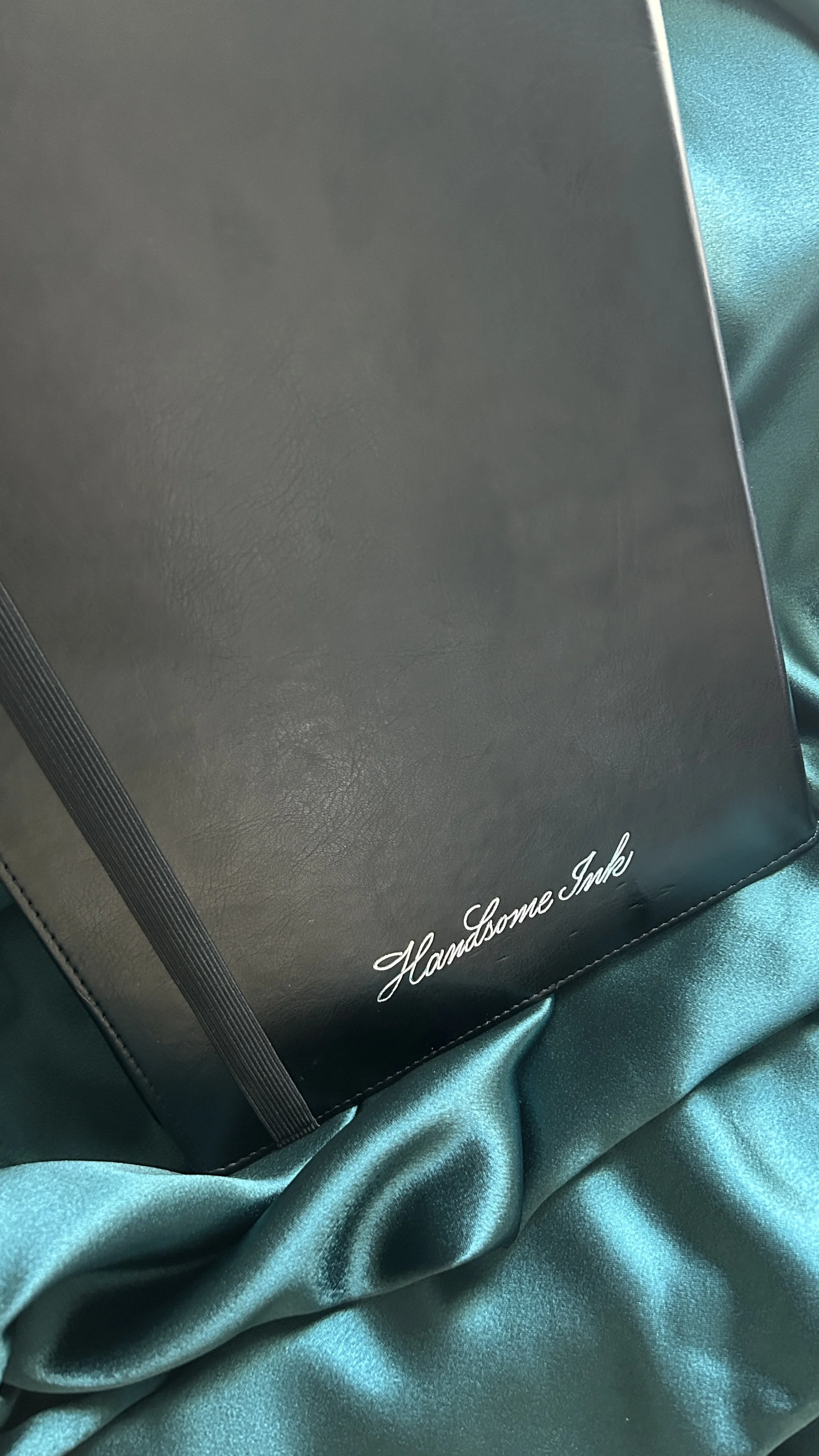 A black notebook with the words 'Handsome Ink' written in white cursive at the bottom right corner, placed on shiny teal fabric.