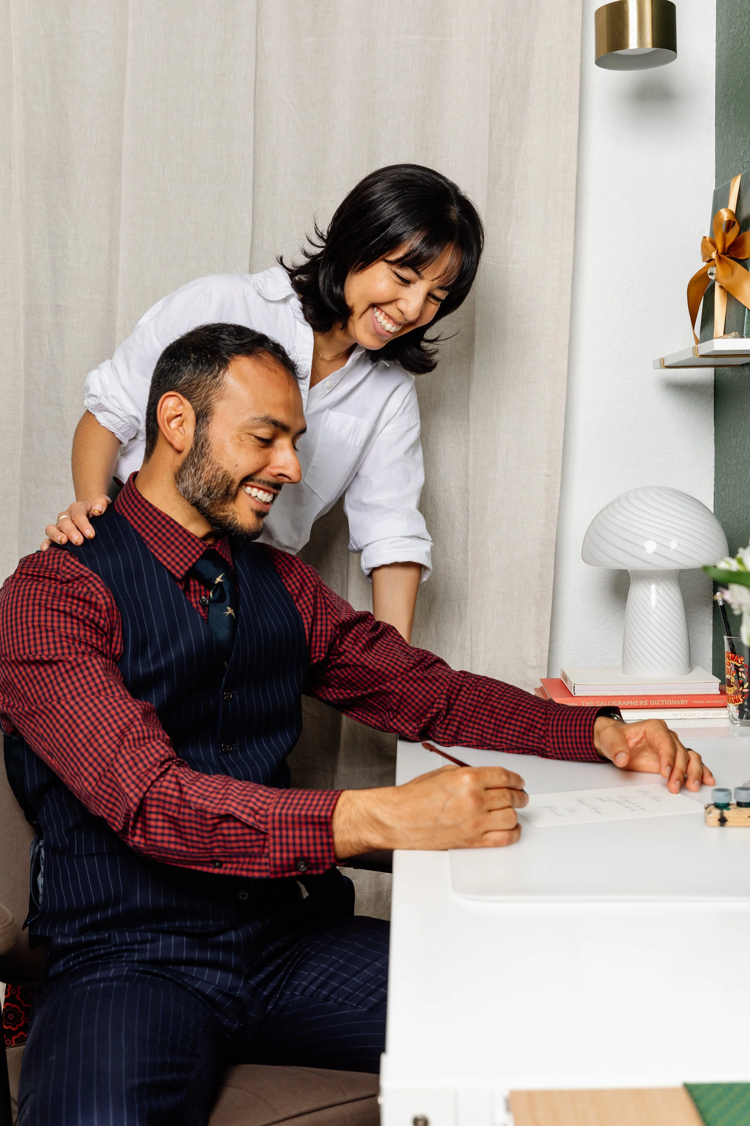 A woman and a man are sitting at a white desk, smiling and looking at papers. The woman is standing behind the man, leaning over and sharing a happy moment. The man is wearing a striped vest and a red checkered shirt. The scene appears to be in a modern, cozy office or home office setting with decorative items on a shelf and a lamp.