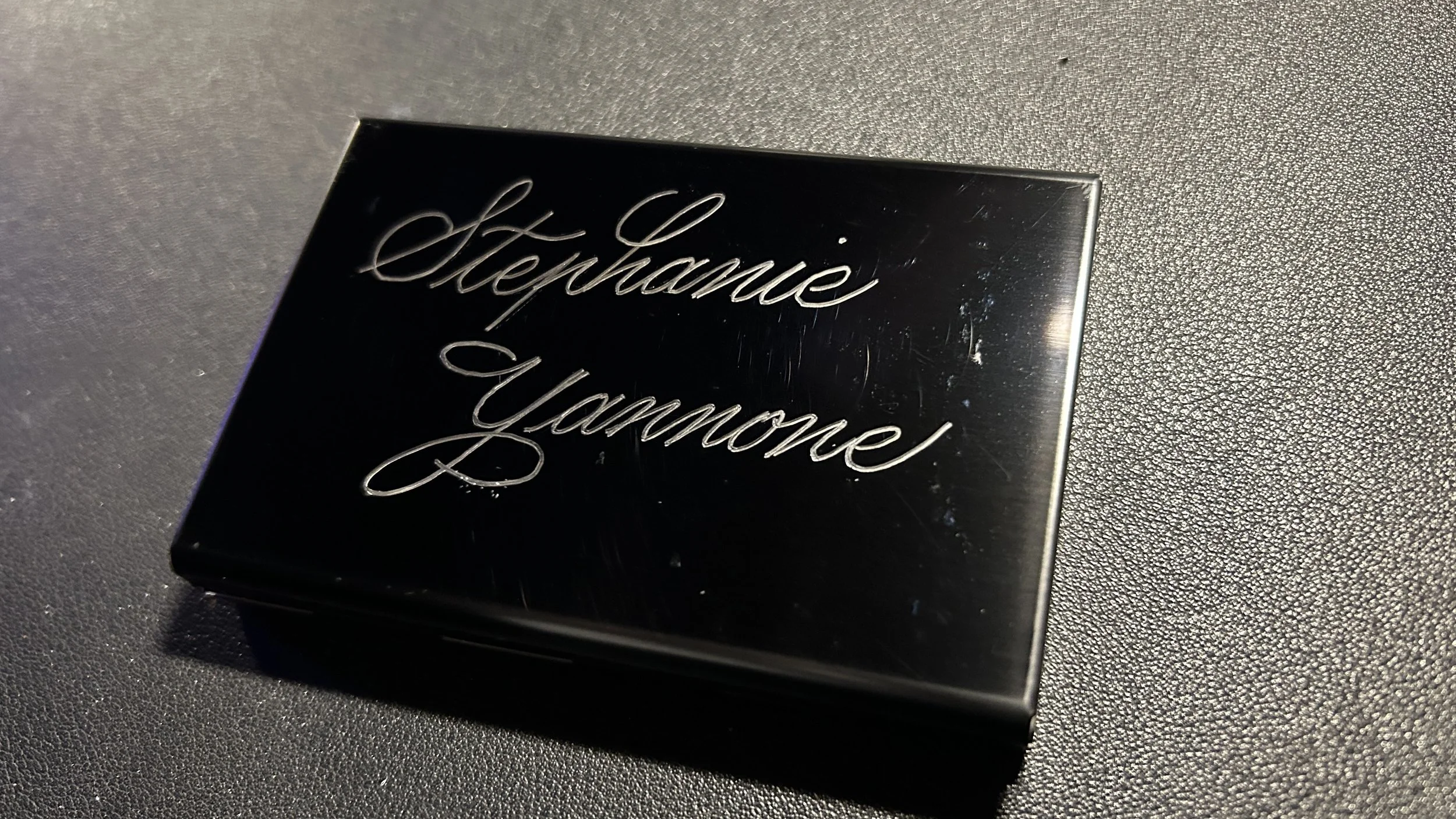 A small black box with silver cursive lettering that reads 'Stephance Yvonne' resting on a textured dark surface.