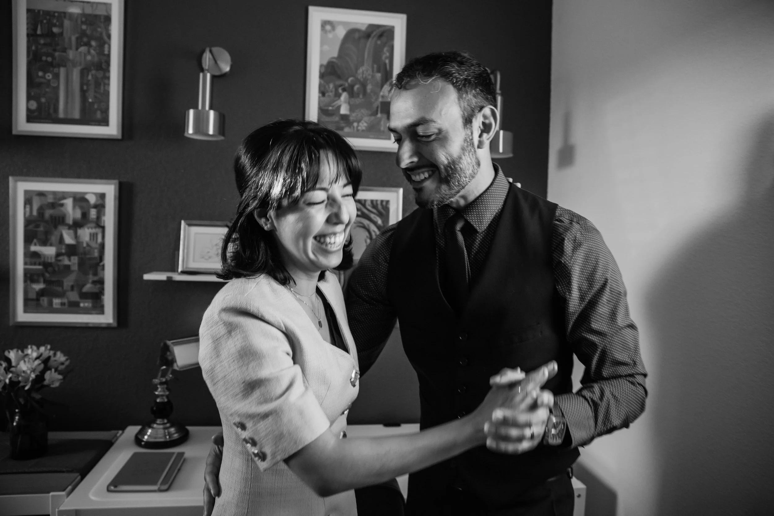 A black and white photo of a man and woman smiling and dancing together in a room with framed artwork on the wall behind them.