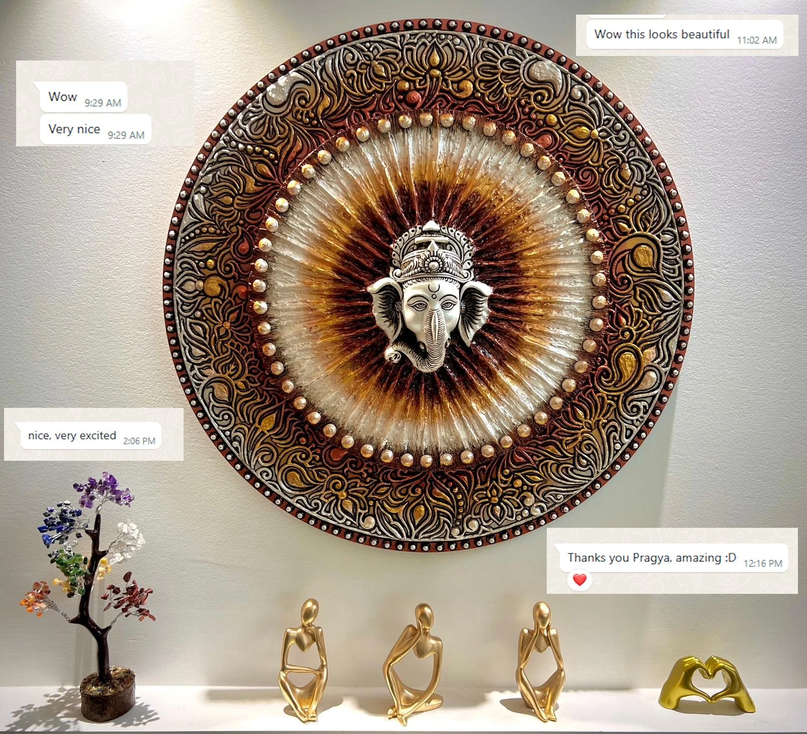 Decorative wall art featuring an ornate circular design with a central Ganesha face. Below are small sculptures of stylized human figures, a colorful tree sculpture, and a gold heart-shaped sculpture.