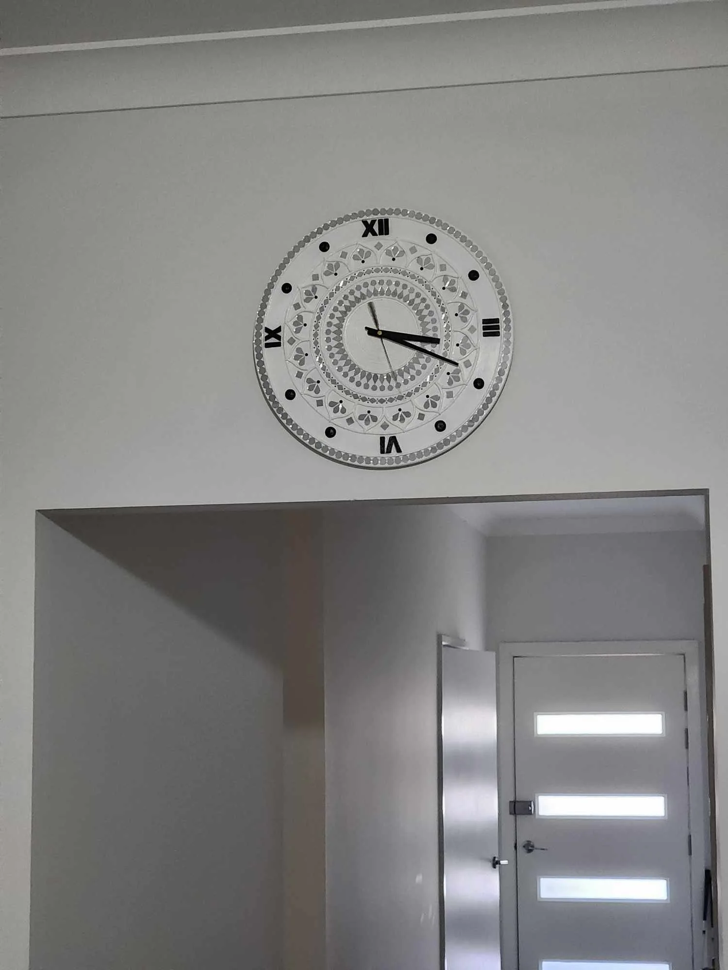 A decorative wall clock with black Roman numerals and intricate patterns hangs above an entrance doorway with a white door featuring horizontal rectangular windows.