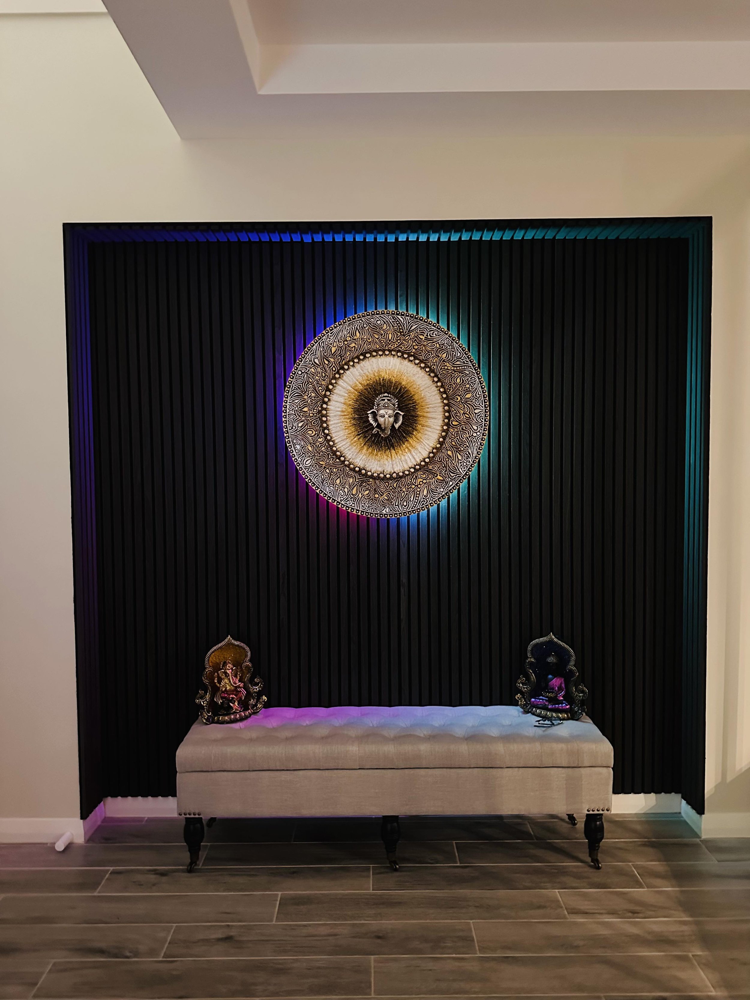 Decorative wall with black vertical slats, a round artwork with a lion's face at the center, illuminated with colorful LED lights, a beige bench with two small Hindu deity statues on it, and a wood-look tile floor.
