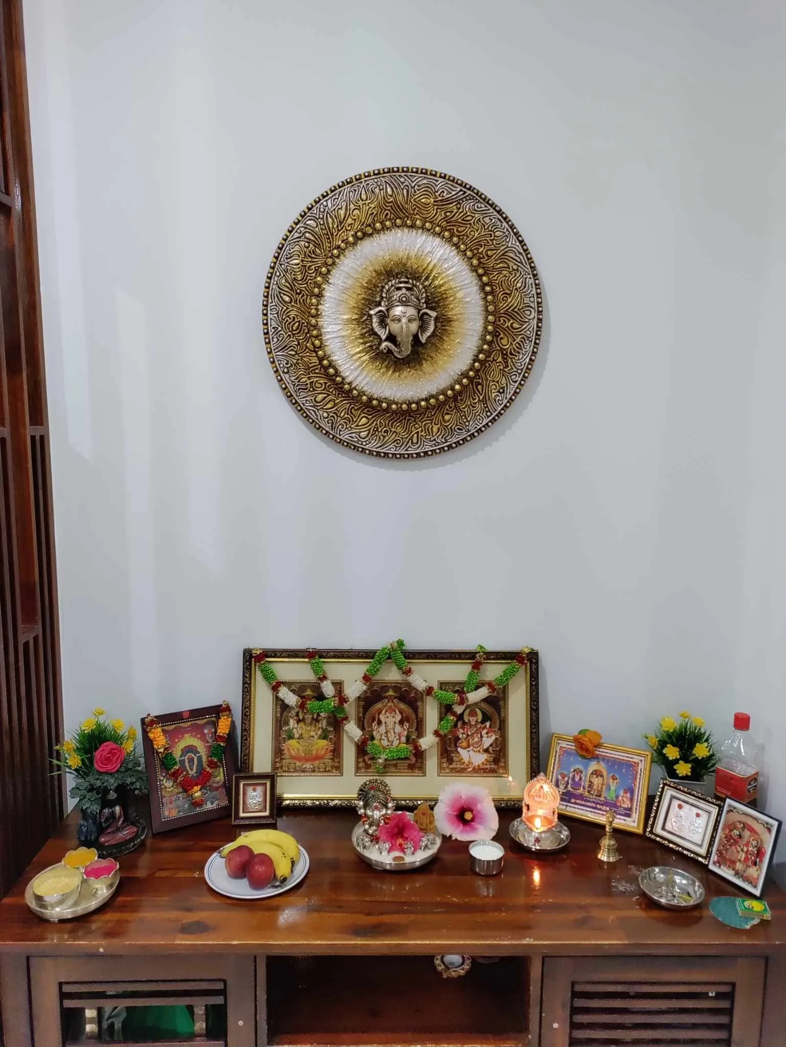 Hindu prayer altar with framed pictures of deities, flower garlands, candles, fruits, and other offerings on a wooden table against a plain wall.