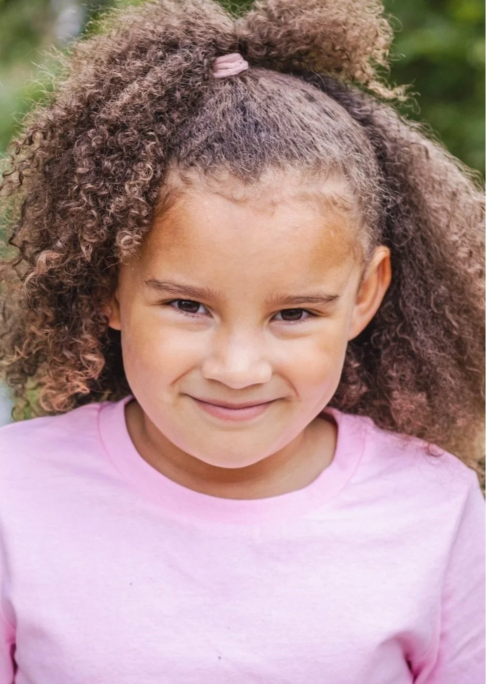 A young girl with curly hair, wearing a pink shirt, smiling outdoors with greenery in the background.