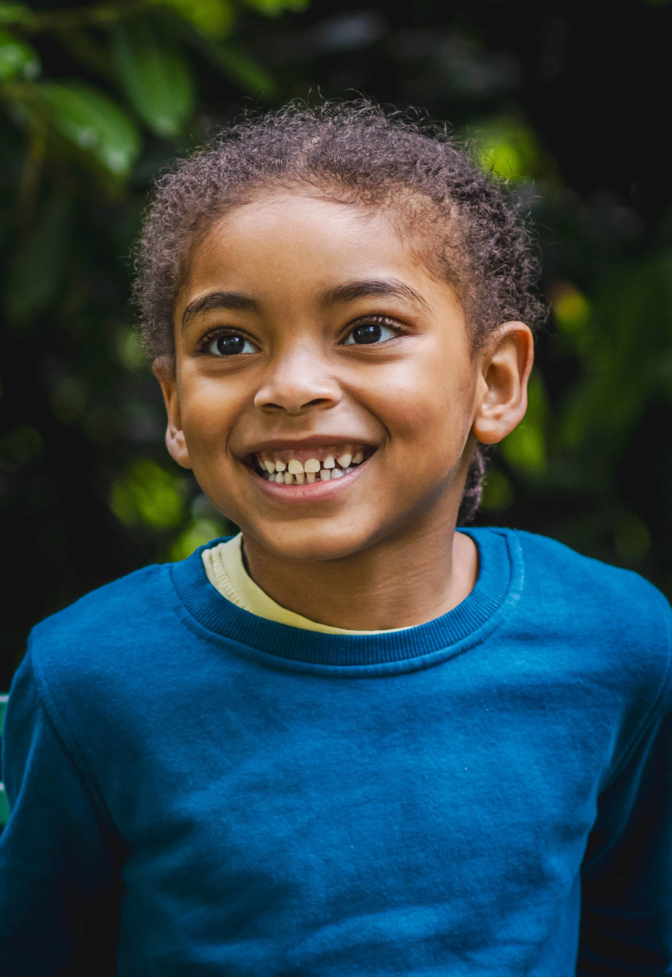 A young boy with curly hair and brown skin, smiling and showing missing teeth, wearing a blue shirt and standing outdoors with green foliage in the background.