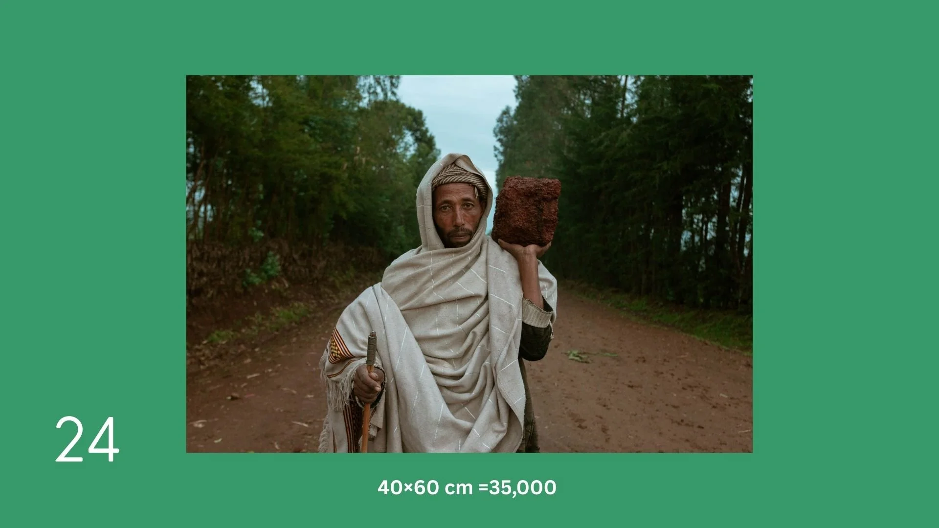 A man dressed in traditional clothing carrying a large brick on his shoulder and holding a walking stick, standing on a dirt path surrounded by trees.