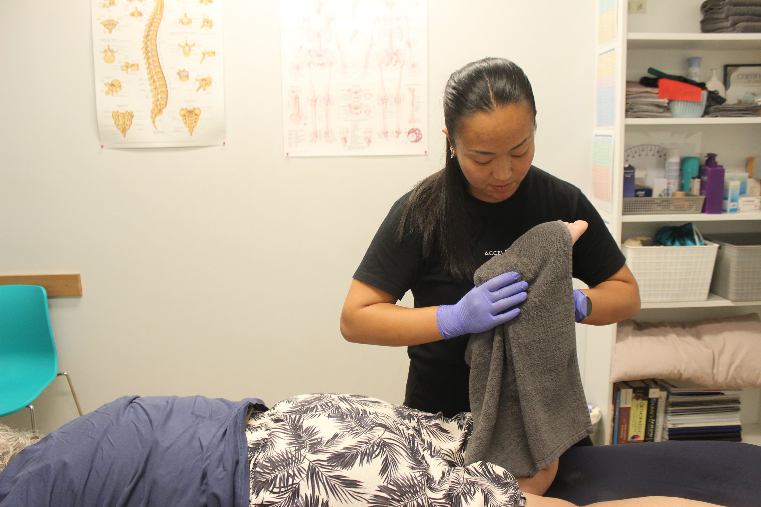 A woman with black hair, wearing a black T-shirt and purple gloves, performing a medical procedure on a person lying on a treatment table. The person on the table has blonde hair and is wearing a printed shirt. The room has medical posters on the wall and shelves with medical supplies.