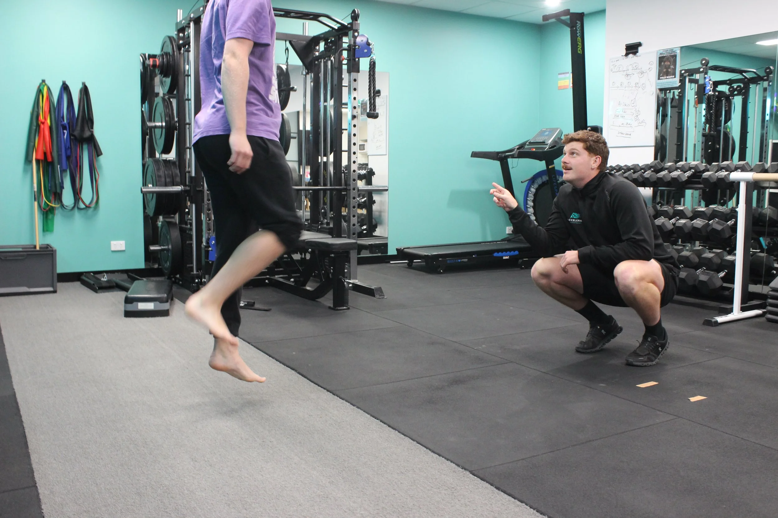A person jumping on a trampoline in a gym while a trainer or coach crouches down, giving instruction. The gym has various workout equipment, including dumbbells and resistance bands.