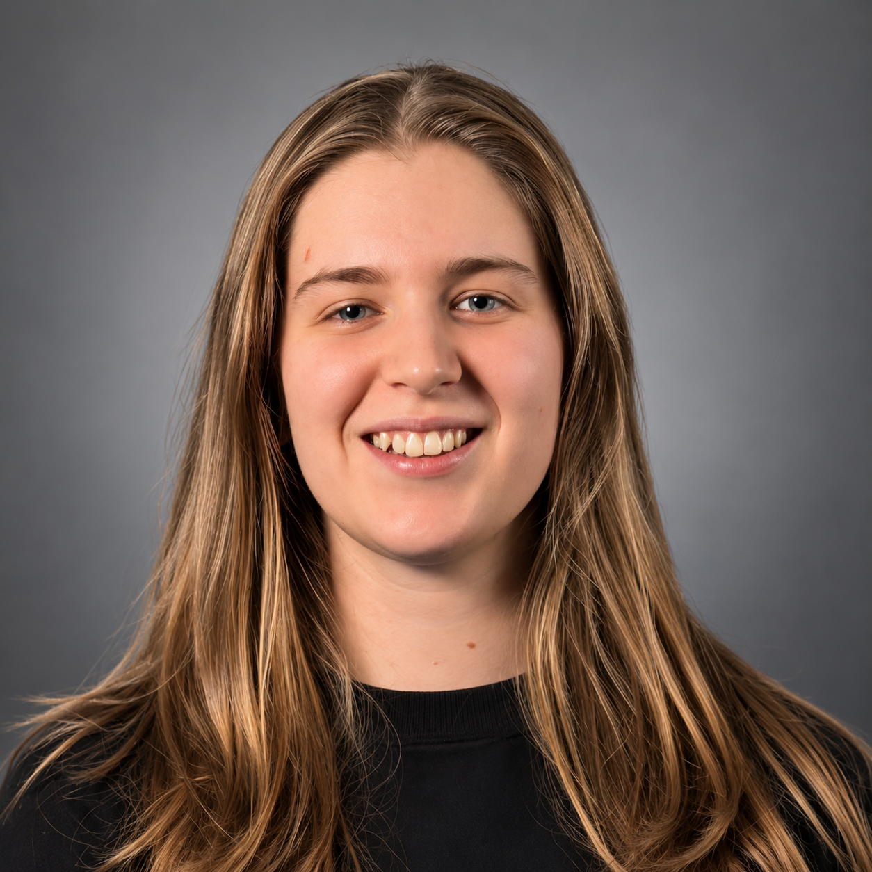 Close-up portrait of a smiling young woman with long wavy blonde hair and blue eyes, wearing a black top against a plain gray background.