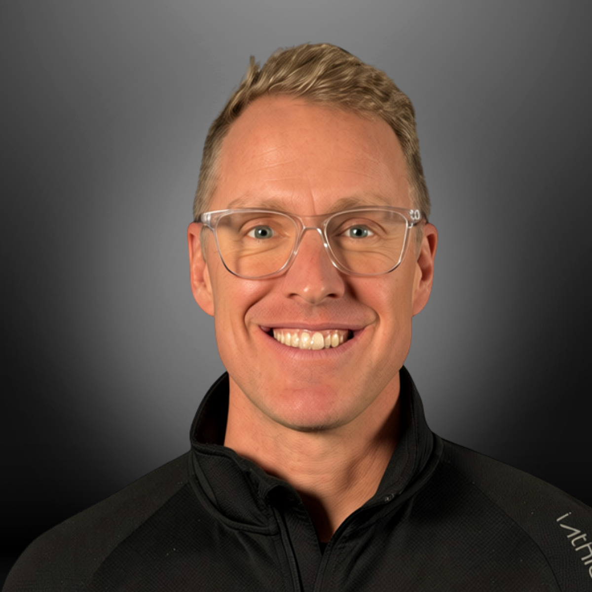A smiling man with blond hair, glasses, and a black athletic jacket against a gray gradient background.
