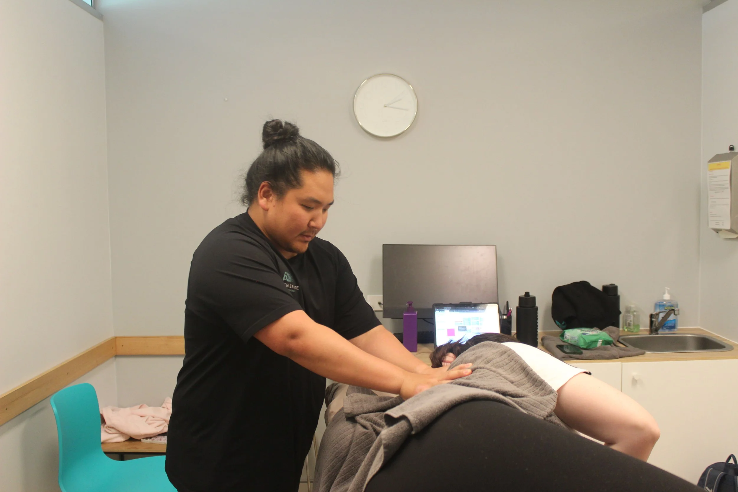 Person receiving a massage in a therapy room with various supplies on the counter, a computer monitor, and a wall clock showing about 10:14.