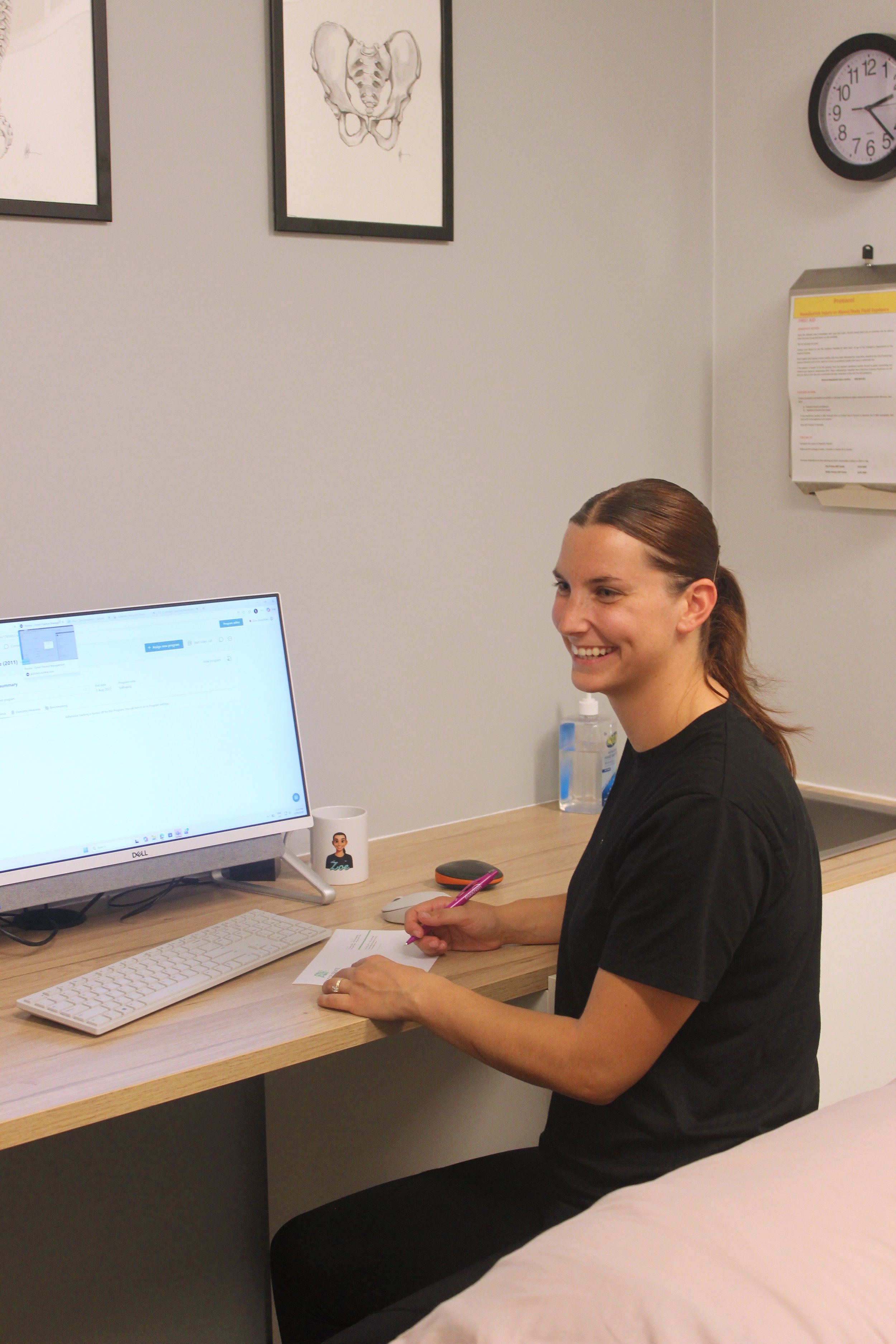 A woman with brown hair tied back, wearing a black shirt, sitting at a desk in an office, smiling while writing on a piece of paper. There is a computer monitor, a white keyboard, a mouse, a mug with a cartoon character on it, a water bottle, and pink eyeglasses on the desk. The background features framed anatomical art of the pelvis and a wall clock.