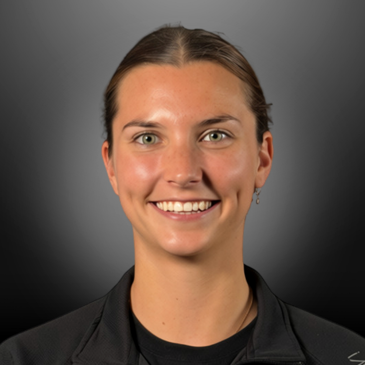 Close-up portrait of a smiling young woman with light skin, brown hair tied back, green eyes, wearing a black top and earring, against a dark gray background.