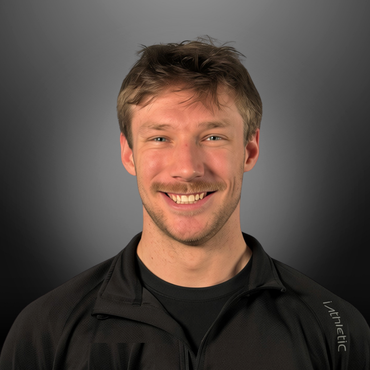 Smiling young man with brown hair and a mustache, wearing a black athletic jacket, against a gray background.
