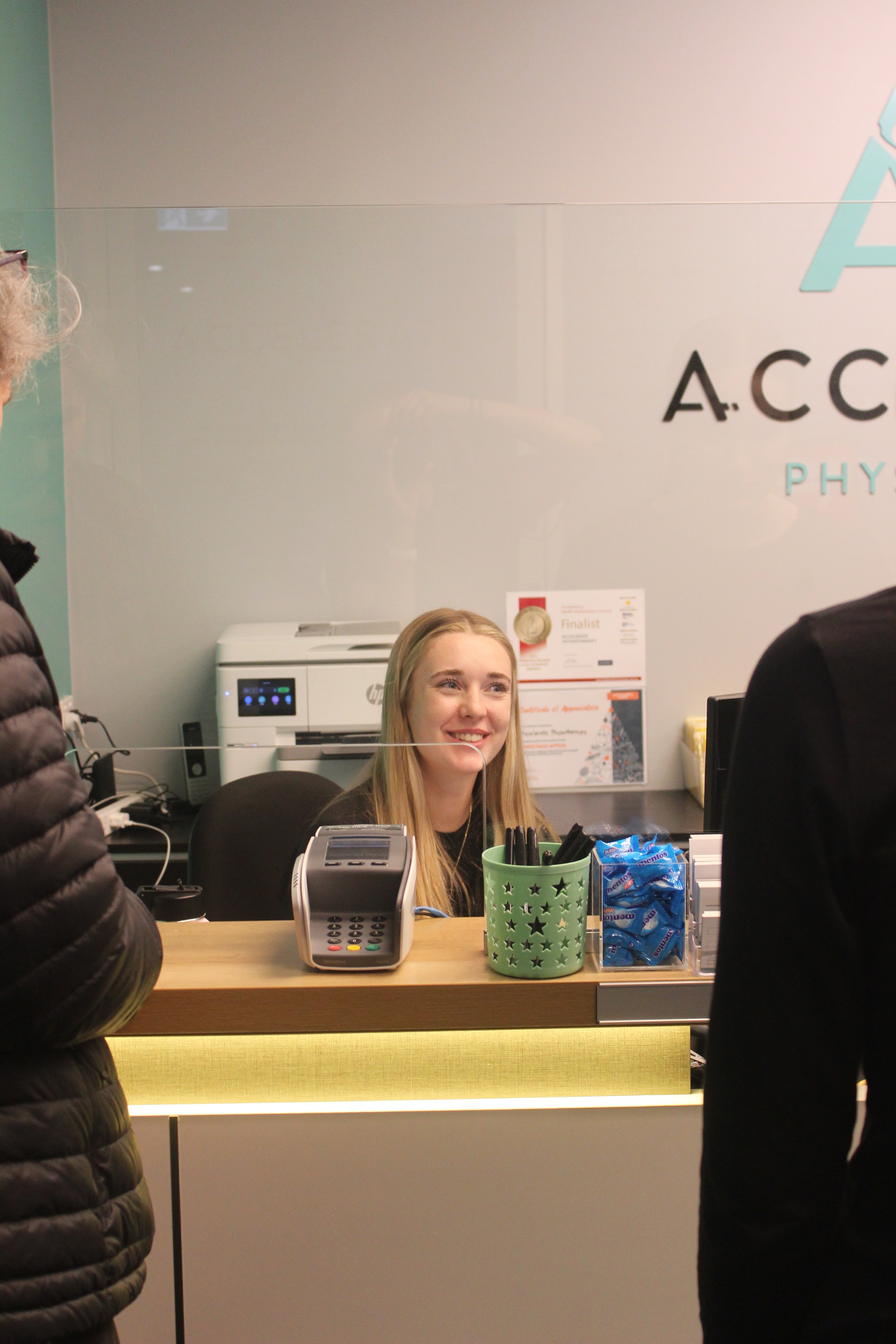 A young woman with blonde hair and a black shirt smiling at a checkout counter. The counter has a card payment terminal, a green cup with pens, and a clear container with blue snack packs. Two people are partially visible on either side, and the background has a sign with the words 'A C...' and 'PH...' and an image of a Christmas tree.