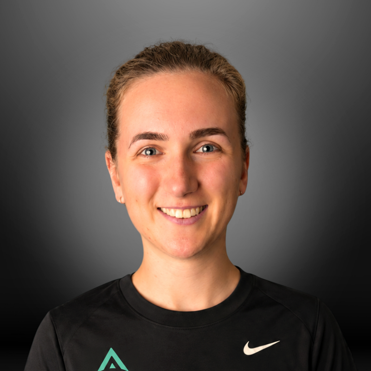 Young woman with short blonde hair, blue eyes, and earrings smiling, wearing a black Nike athletic shirt against a gray background.