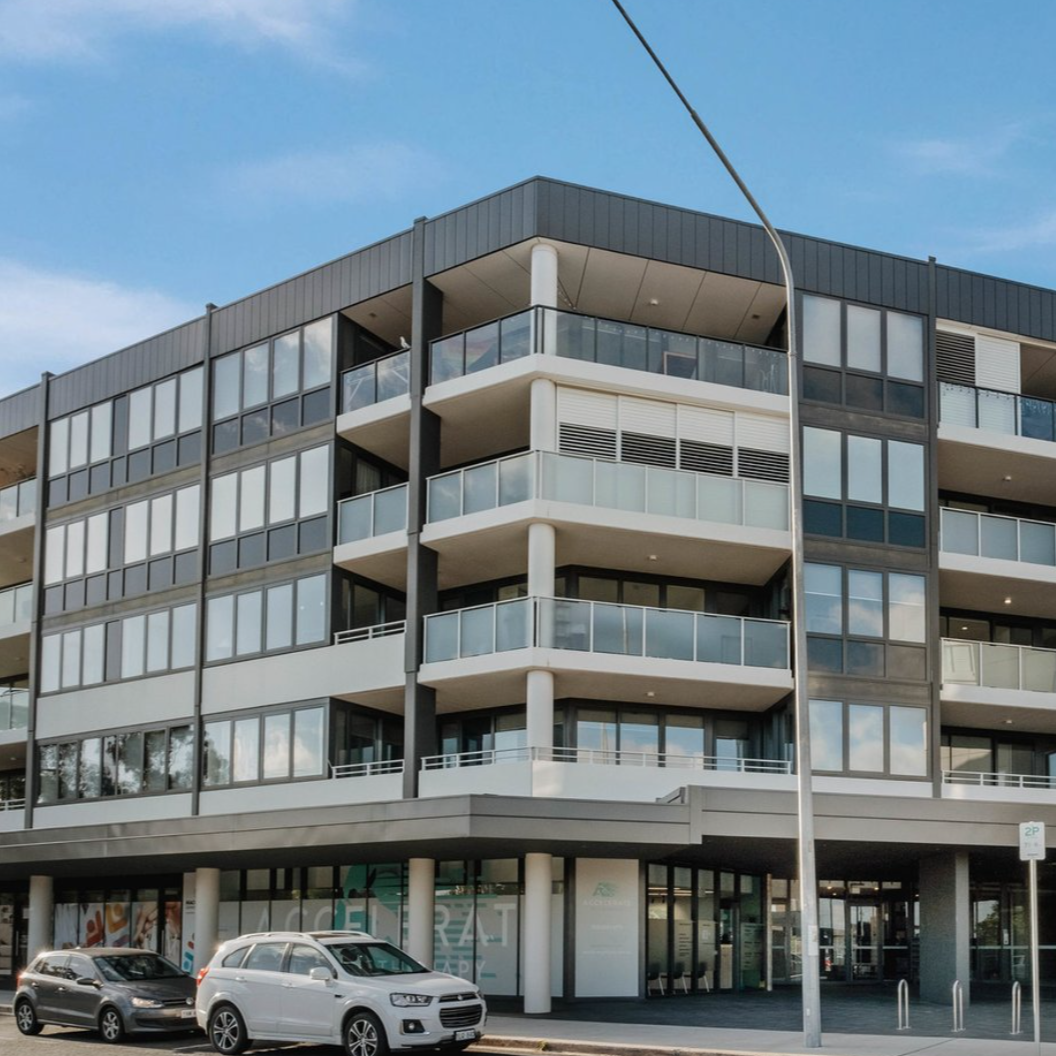 Modern four-story apartment building with glass balconies and large windows, with cars parked in front and a streetlamp nearby.
