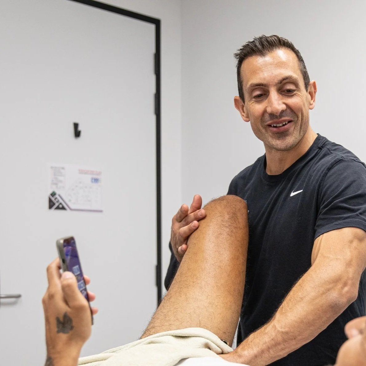 A man giving a leg massage to another person while smiling. The person receiving the massage is lying down. The setting appears to be a health or wellness clinic.