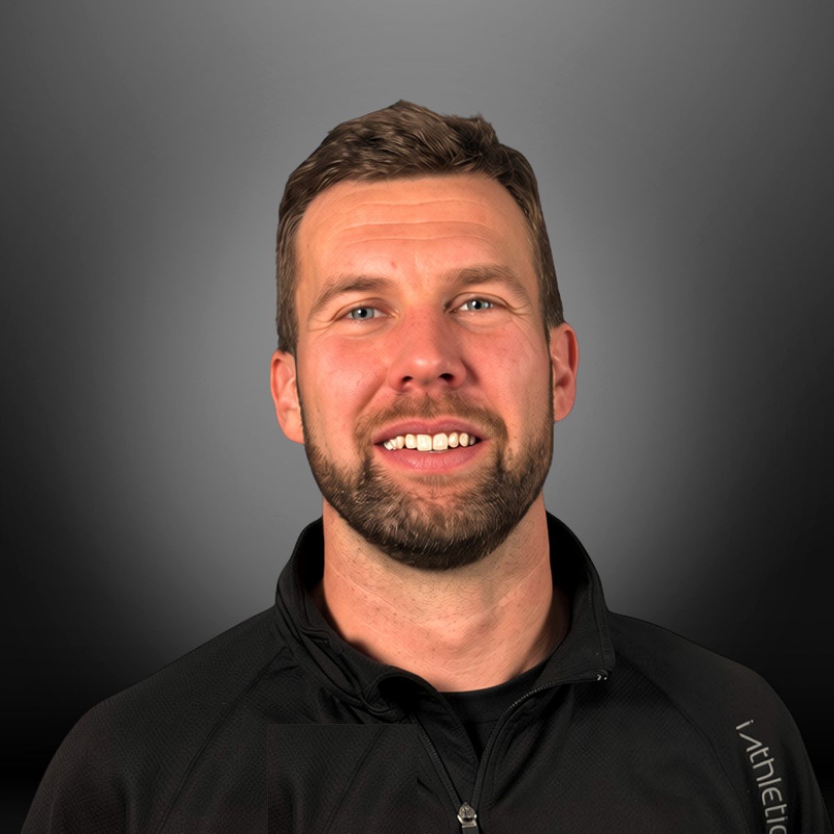 A man with short brown hair, a beard, and blue eyes, smiling, wearing a black athletic jacket with a high collar against a gray background.