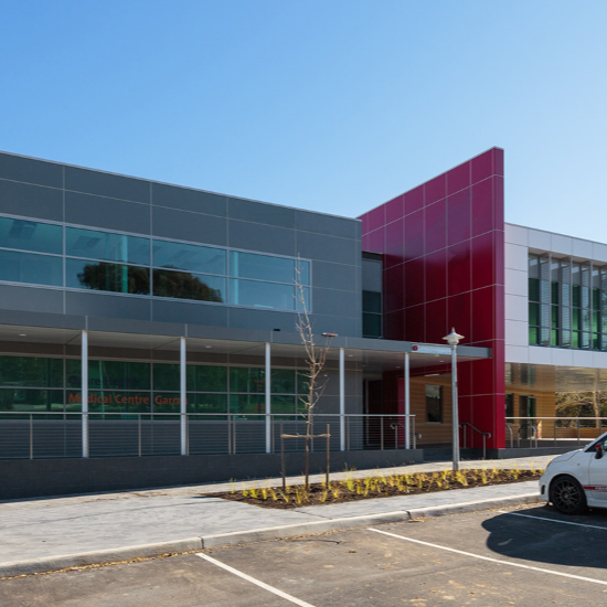 Modern medical center building with red, gray, and green exterior panels and large glass windows, parking lot in front, and clear blue sky.