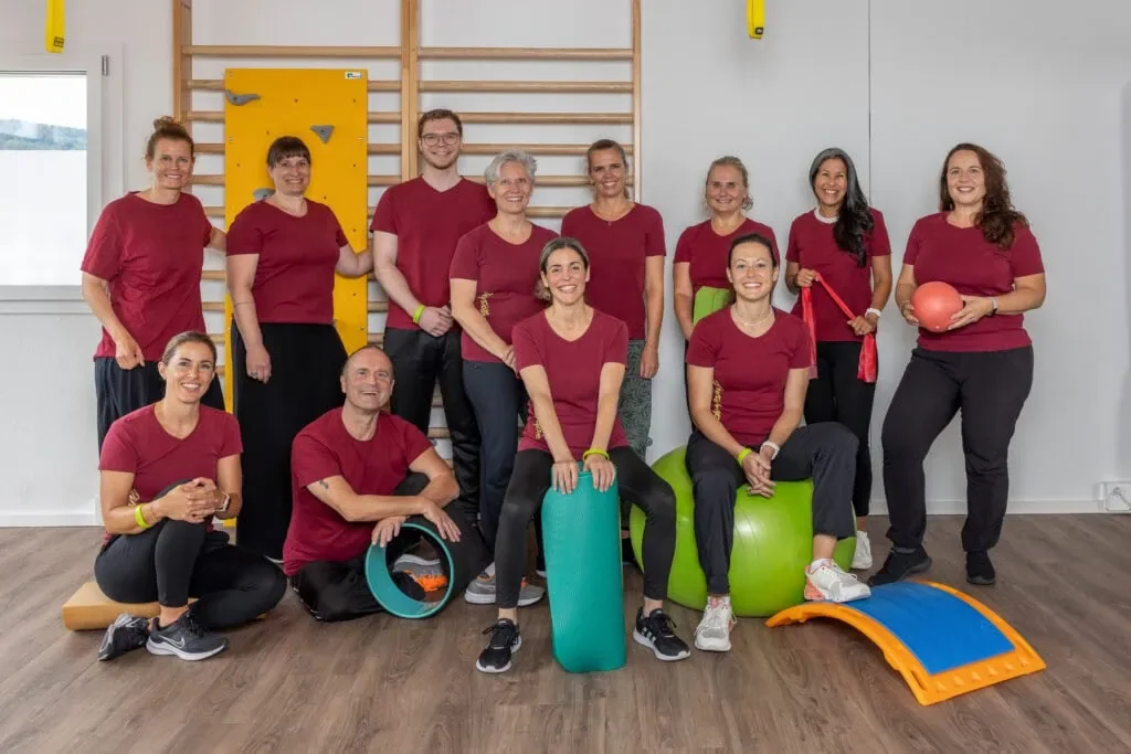 Eine Gruppe von zehn Menschen, die in einem Fitnessraum oder Reha-Zentrum trainieren, alle tragen rote T-Shirts. Manche sitzen auf verschiedenen Fitnessgeräten, andere stehen hinter ihnen macht keiner Übungen. Im Hintergrund eine Wand mit einer Kletterwand und einem Holz Reck. Die Gruppe wirkt fröhlich und motiviert.