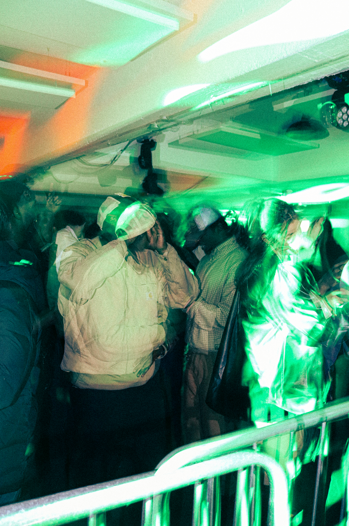 People standing close together indoors with colorful green and orange lighting, some wearing jackets and hats, possibly at a social event or party.