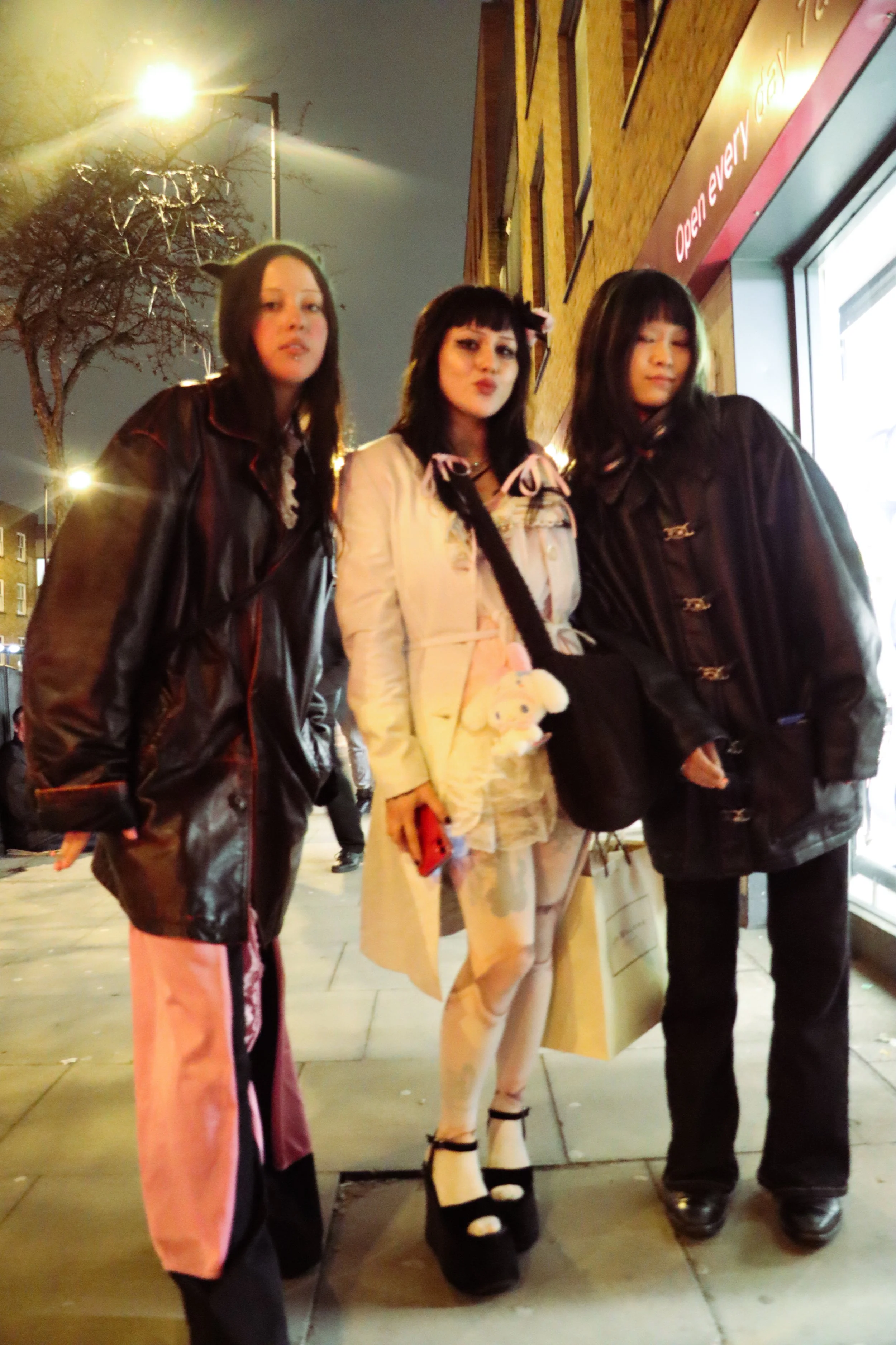 Three young women standing outside at night near a storefront with a sign that says 'Open Everything.' They are dressed in fashionable clothing, with two wearing black jackets and one in a beige coat, and are posing for the photo, some with winks and