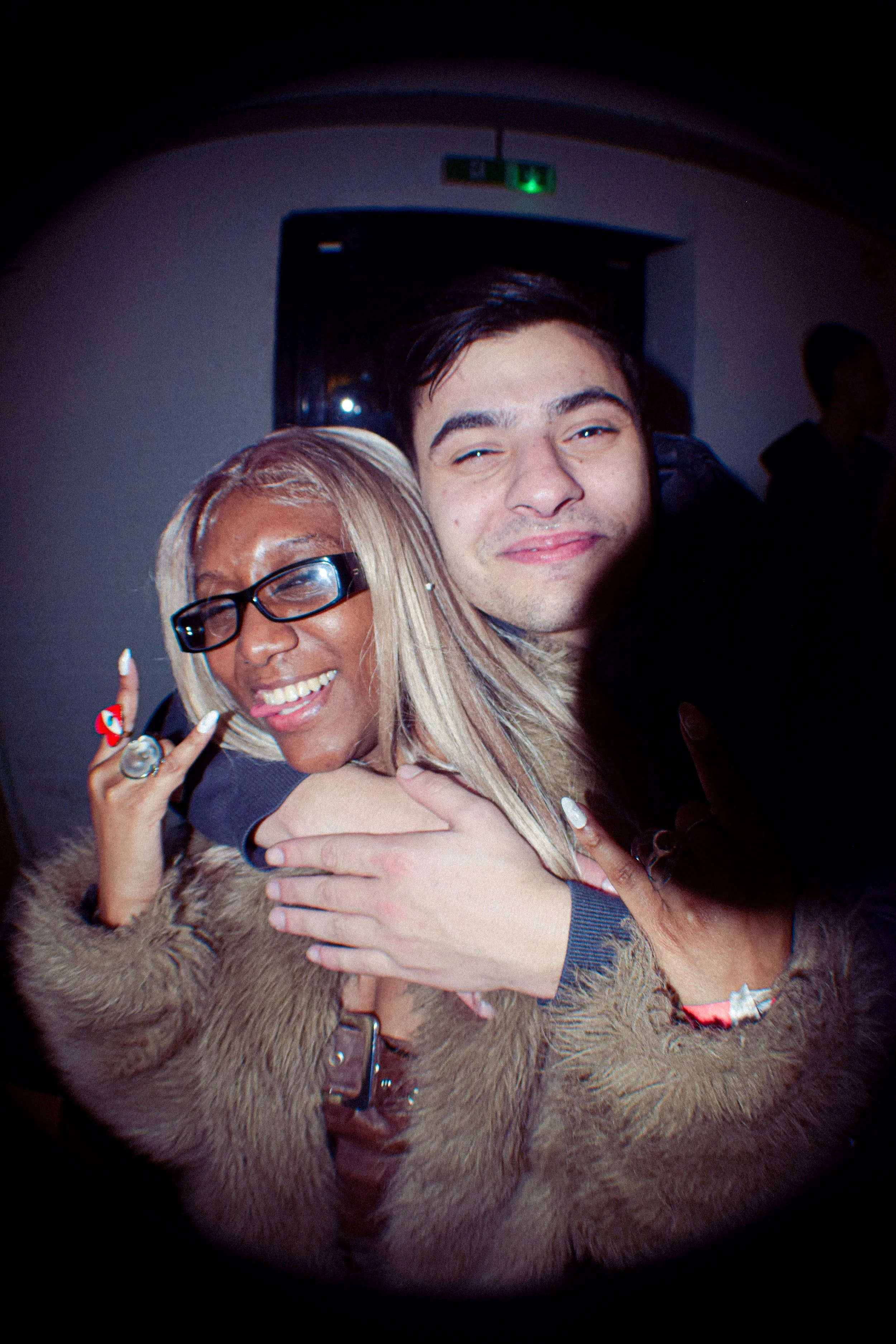 Two people embracing, smiling, and showing friendly gestures, with a dark background and a faint green exit sign above.