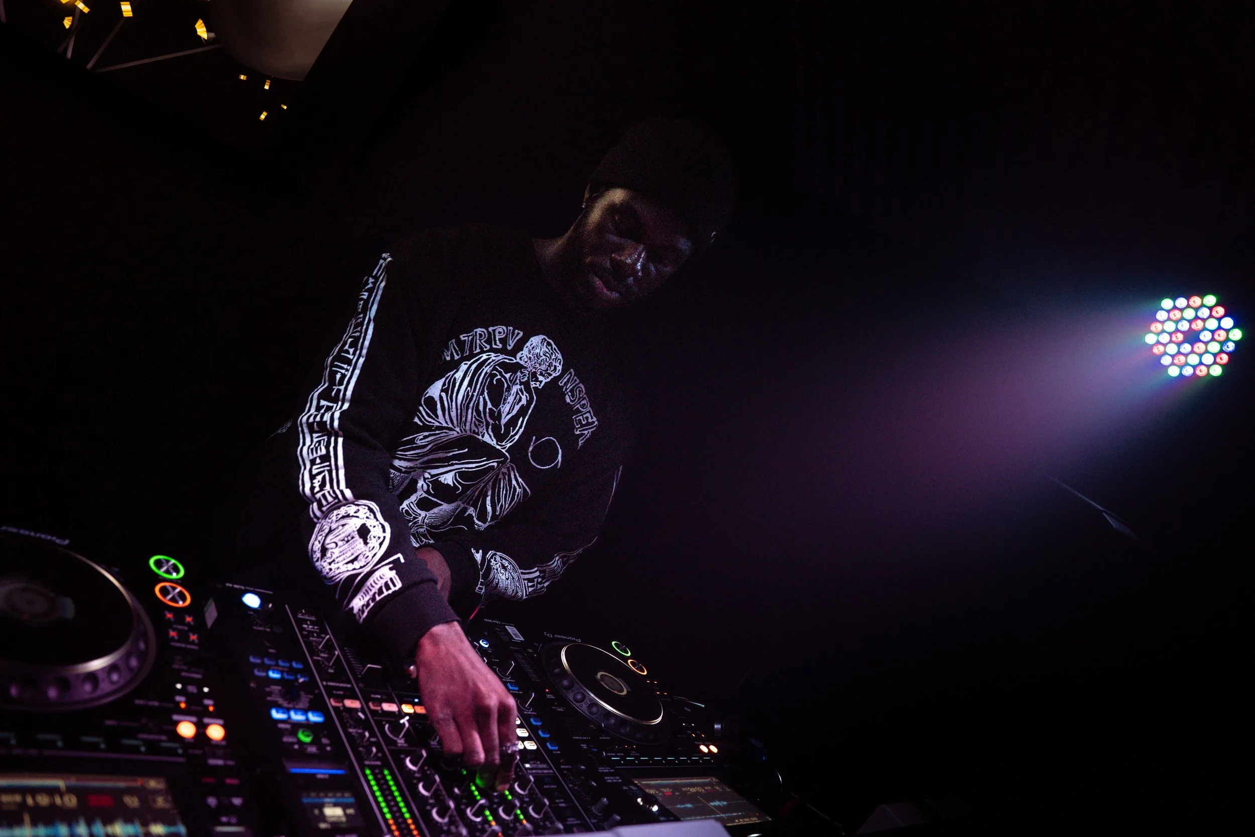 DJ performing in a dark setting, wearing a black hoodie with a graphic design, surrounded by colorful DJ equipment and illuminated by stage lights.
