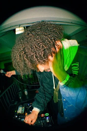DJ with curly hair performing at a club or party, using a mixer and wearing a green jacket.