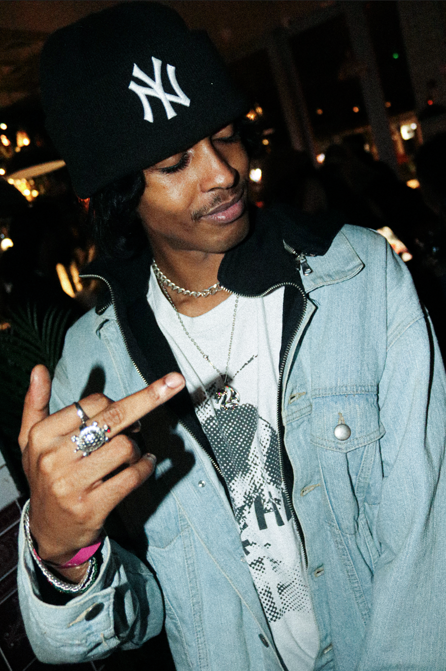 A man wearing a black New York Yankees cap, a silver chain necklace, a graphic t-shirt, and a light denim jacket is pointing at himself with his middle finger. He has medium-length black hair and is in a crowded indoor setting with dim lighting.