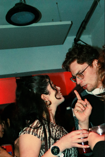 A man and woman engaging in conversation at a social gathering or party, with the woman smiling and holding a drink, and the man gesturing with his finger.