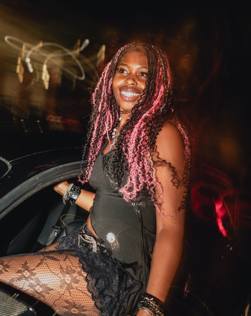 A smiling woman with pink and black braided hair, wearing a black dress with lace stockings, sitting on a car door at night with colorful blurred lights in the background.