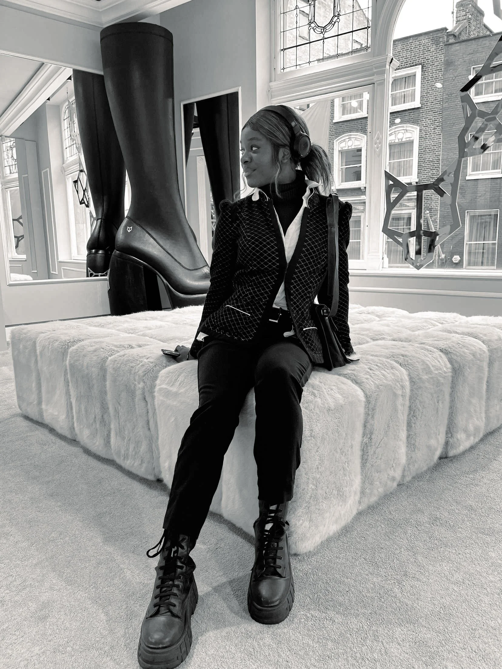 A young woman in black clothing, wearing headphones, is sitting on a plush, oval-shaped bed in a well-lit room with large windows. Behind her, oversized black boots are displayed vertically. The room has ornate moldings and a decorative stained glass window outside.