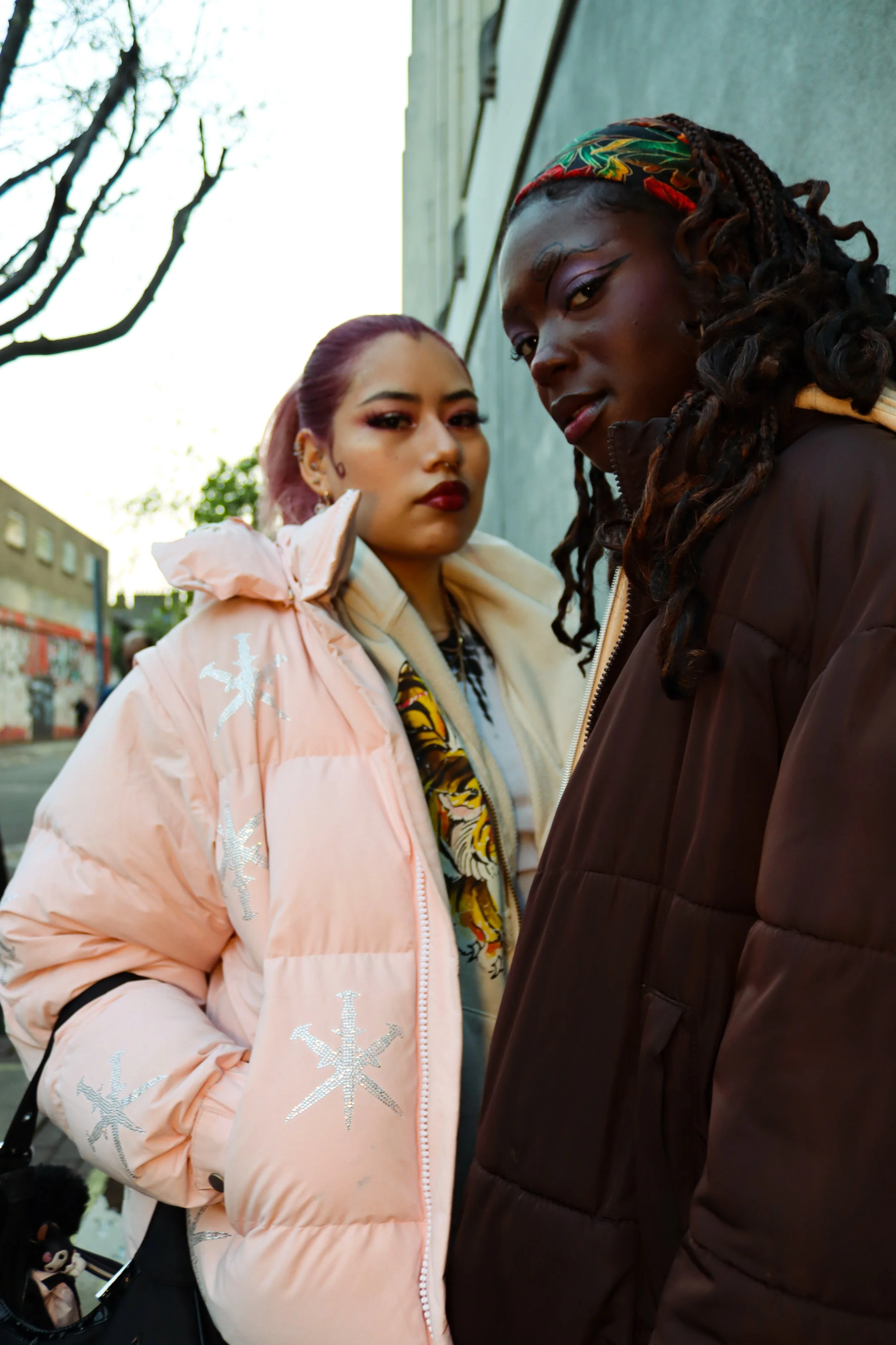 Two women standing close together outdoors, one wearing a pink jacket with snowflake patterns and the other wearing a dark brown jacket, looking at the camera.
