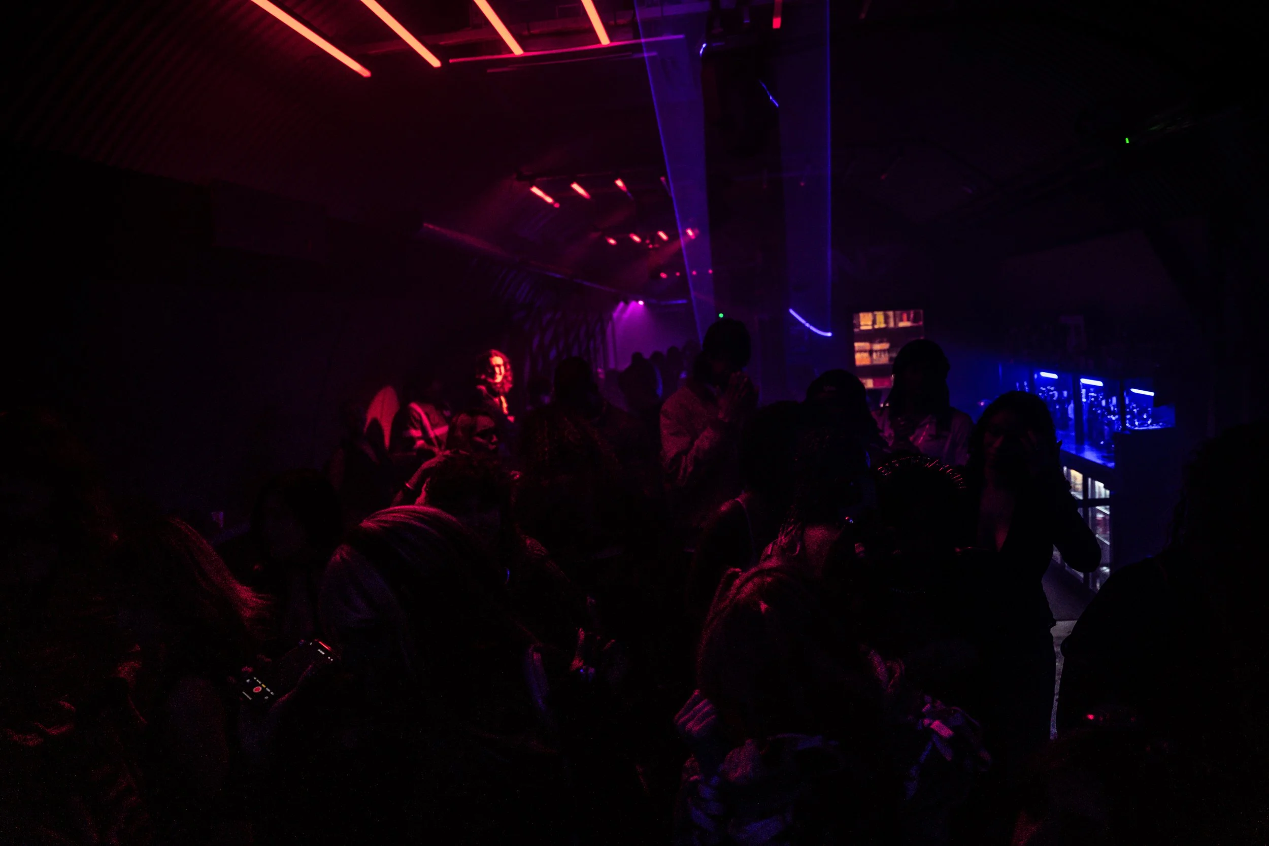 A dark nightclub with people dancing and socializing under colorful neon and laser lights, including red, purple, and blue beams.