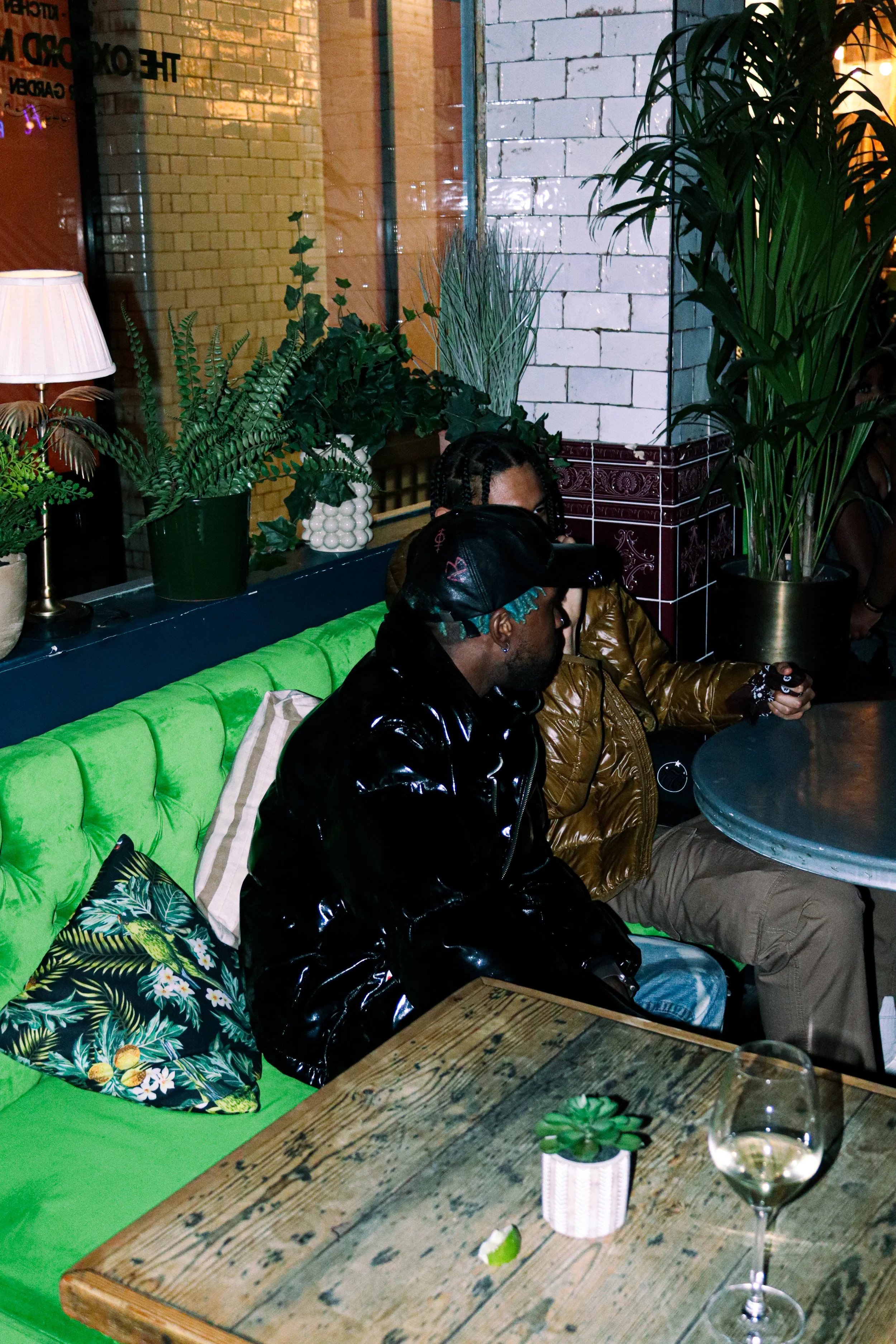 A cozy indoor scene with a rustic wooden table, a green cushioned bench, and a variety of potted plants. There are two people sitting, one with braided hair and the other wearing a leather jacket, partially visible in the background. A wine glass wit