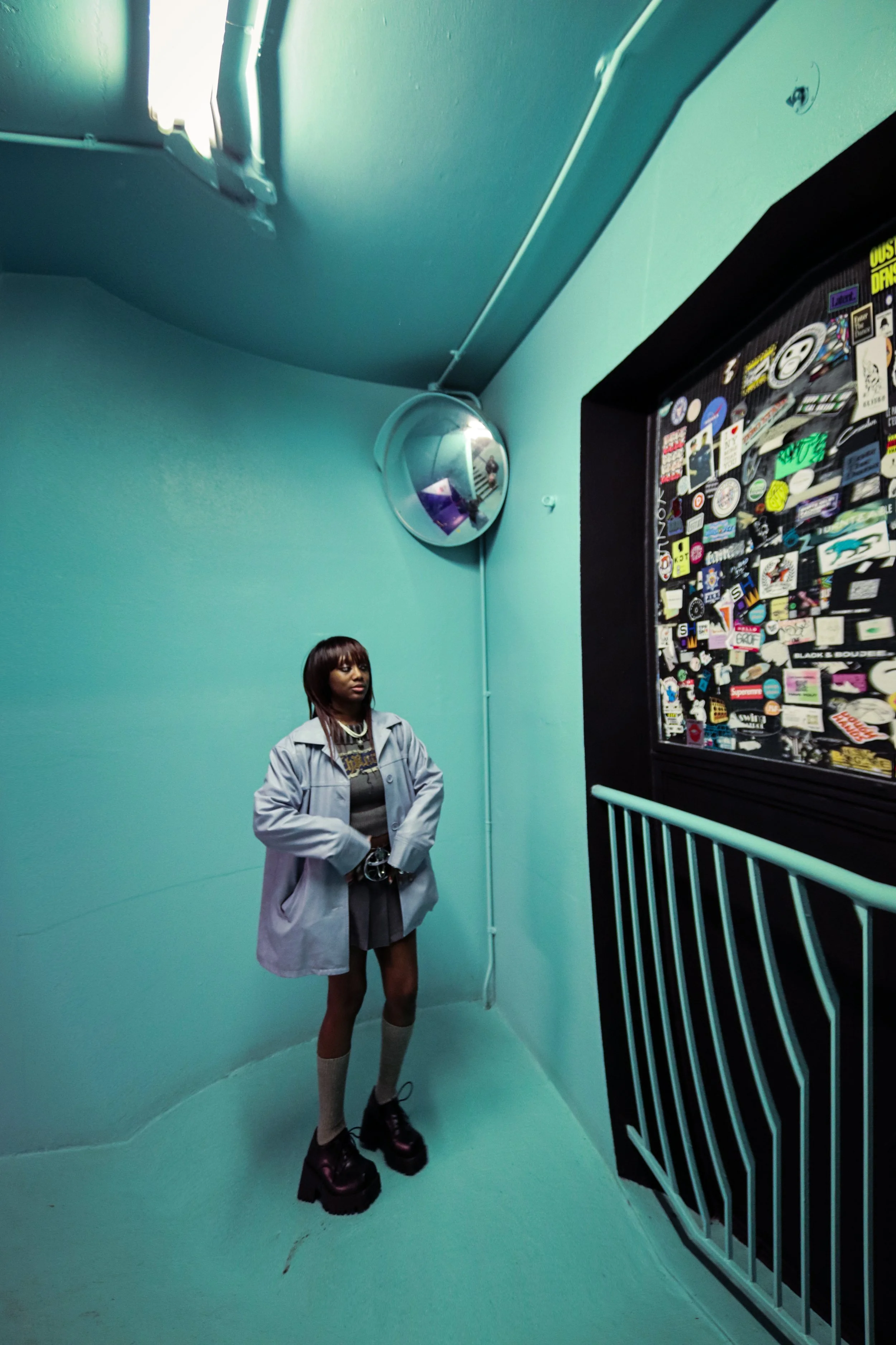 A woman in a light gray coat, black platform boots, and gray socks standing in a teal room near a large sticker-covered black surface. She has shoulder-length hair and appears to be looking slightly to the side.