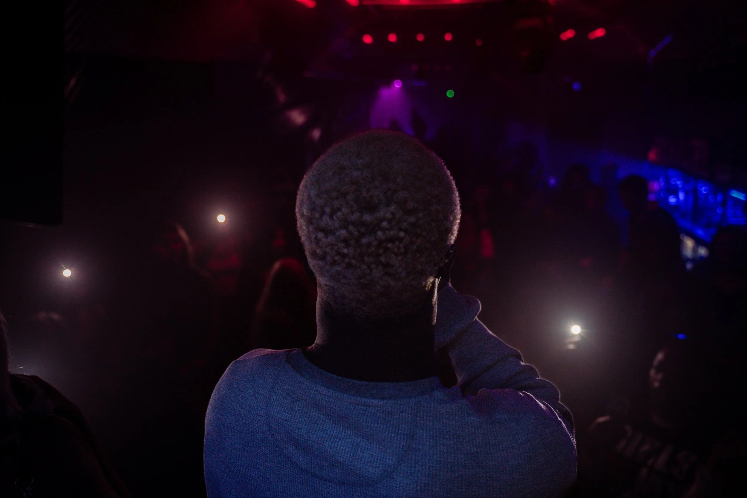A person wearing a gray knit cap and white shirt is seen in silhouette against colorful stage lighting, possibly at a concert or nightclub.
